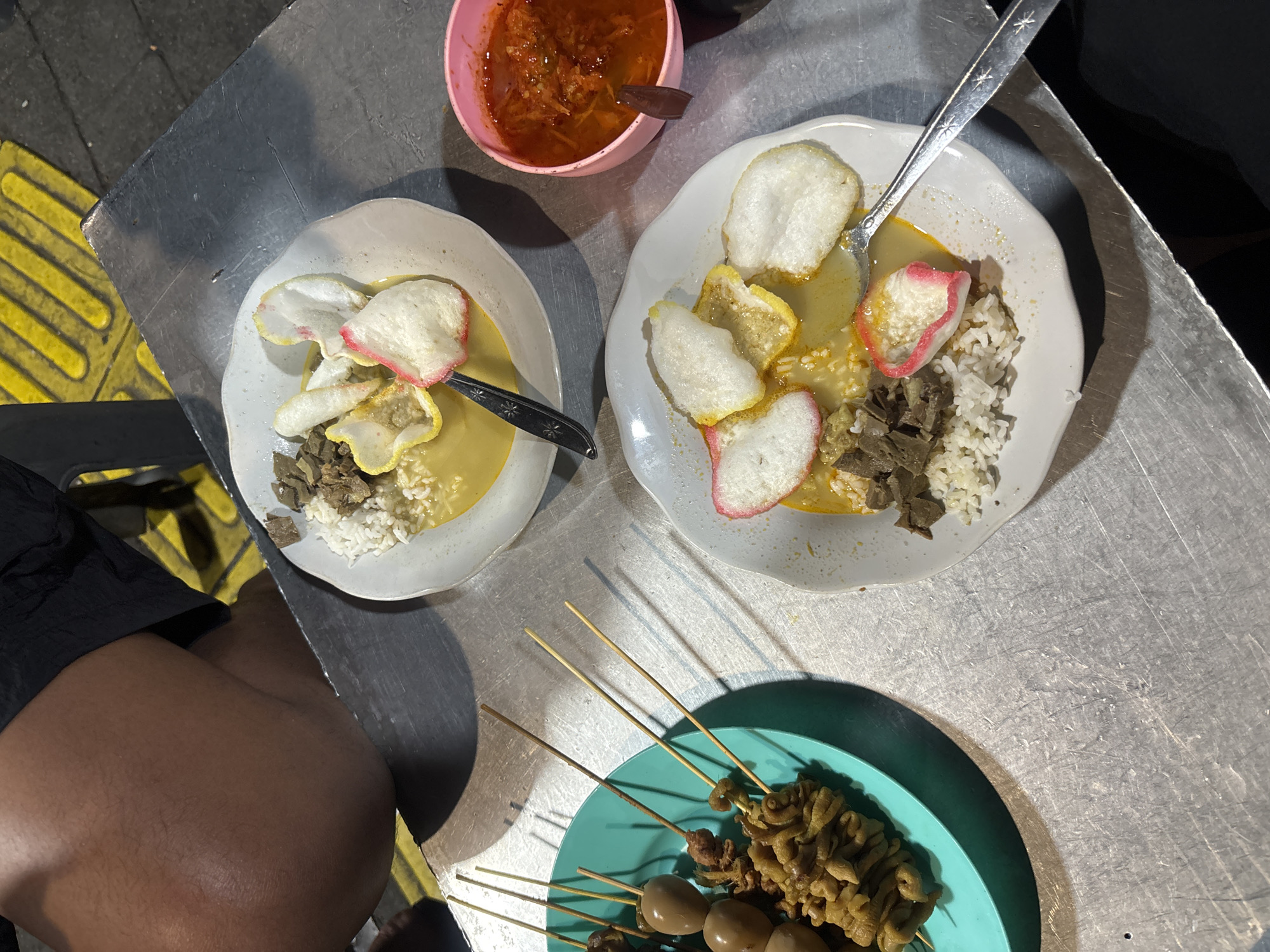 The image shows a casual street food scene. The setting is likely an outdoor dining area with a metallic table, indicated by the reflective surface. The presence of a yellow tactile paving marker in the lower-left suggests the location might be near a pedestrian crossing.
There are two plates of food featuring rice, meat, and crackers in a yellow sauce. Each plate has a spoon resting on it. A pink bowl of chili sauce is also visible. Additionally, a turquoise plate contains skewers of what appears to be quail eggs and chicken intestines.
Based on the food and the overall scene, the location can be inferred to be in South Jakarta, Indonesia, where street food is common. A person's thighs are visible to the left of the shot.