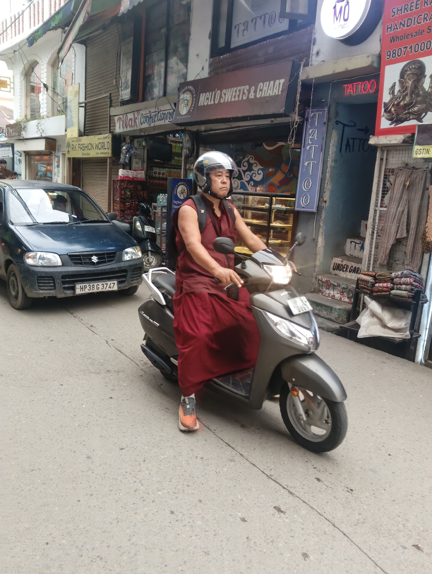 In Dharamkot, India, a man wearing a red robe and a silver helmet rides a gray scooter down a paved street. The man's left arm is visible, resting on the scooter's handlebars. Behind him, a blue car with the license plate "HP38 G 3747" is parked. The street is lined with shops, one of which has a sign that reads "TILAK Confectionery." Another shop features a sign with red lettering that says "TATTOO." The buildings on either side of the street are multi-story structures with various signs and storefronts, suggesting a commercial area. The overall scene depicts a moment in a bustling street in an Indian town.