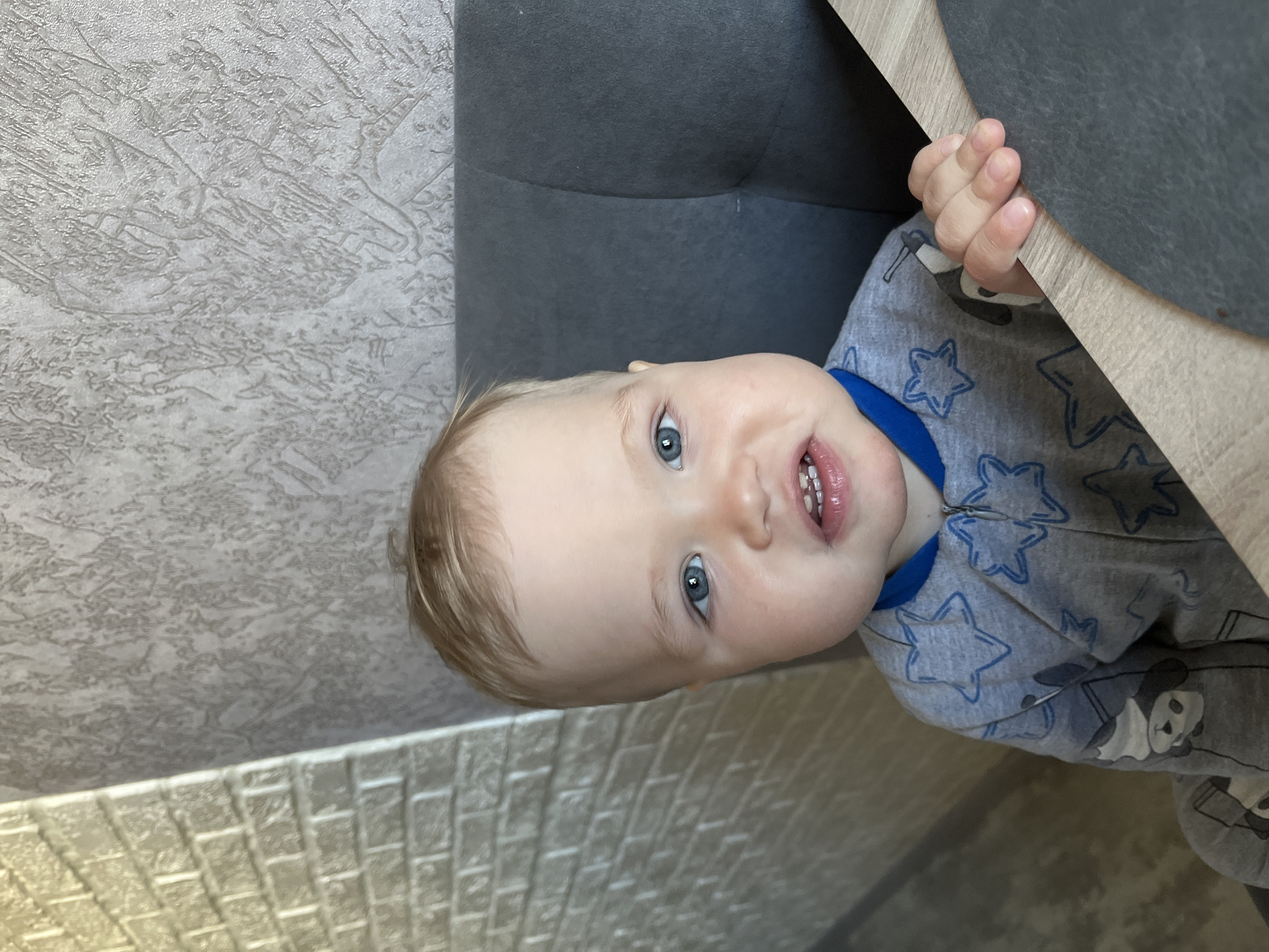 A fair-skinned baby with blonde hair and blue eyes looks upward, partially obscured by furniture and architectural details. The baby is wearing a gray pajama top with blue stars and panda bear graphics. The hand of the baby is holding onto the edge of a light-colored table or counter. The background includes a textured wall and a dark gray upholstered piece of furniture, possibly a sofa or chair. The location is in an interior setting, but no specific landmark or feature indicates that the photograph was taken in Chaikovsky, Russia.
