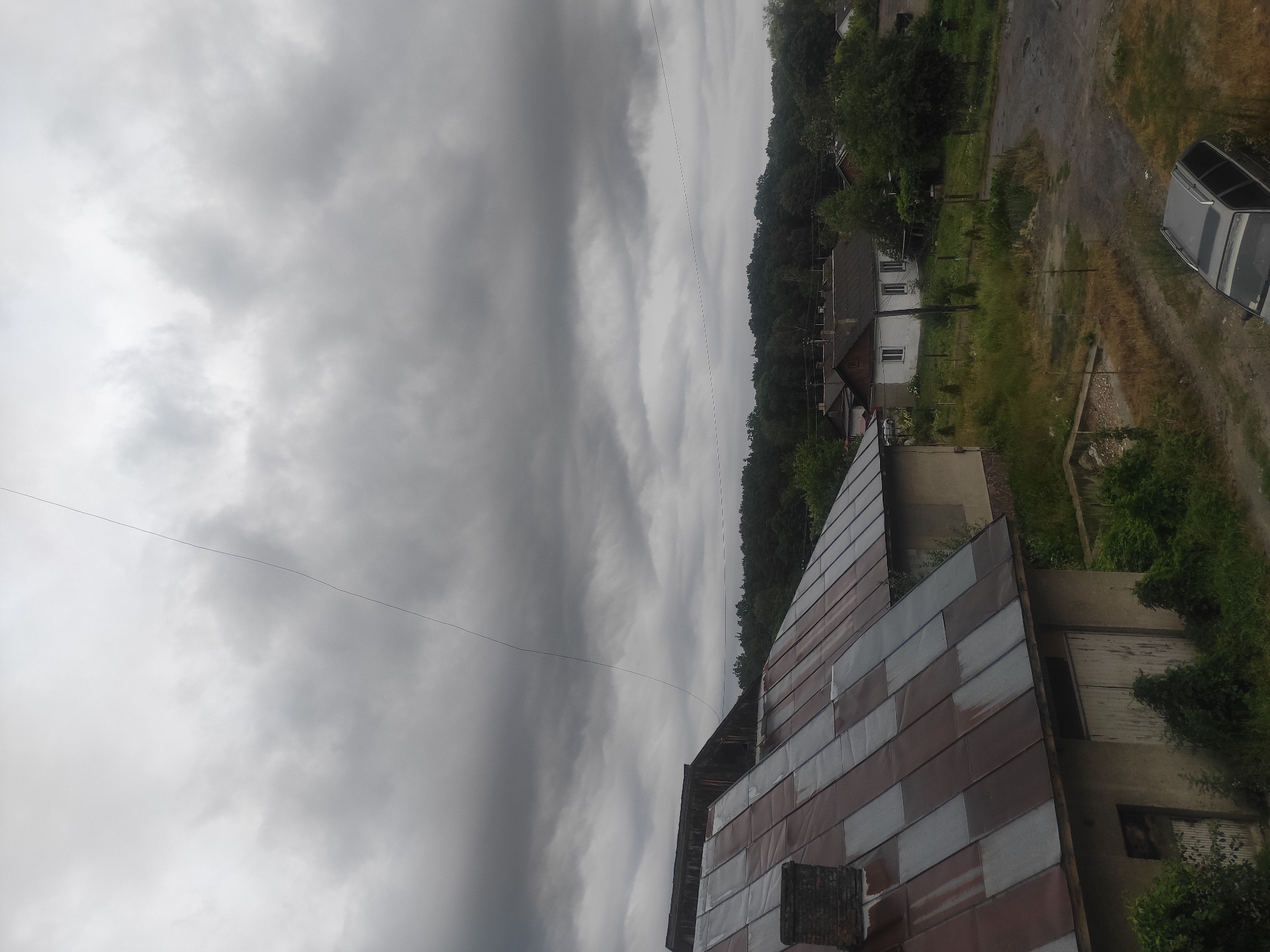 A gray, overcast sky looms over a rural scene in Kostopil, Ukraine. In the foreground, the corrugated metal roof of a building is visible, with a brick chimney on its left. To the right, a narrow, dark line stretches across the sky. Further in the background, several small houses with pitched roofs are scattered among trees and overgrown vegetation. A dirt road winds through the scene, with a dark-colored station wagon parked on it. The overall impression is one of a quiet, possibly somber, day in a village.