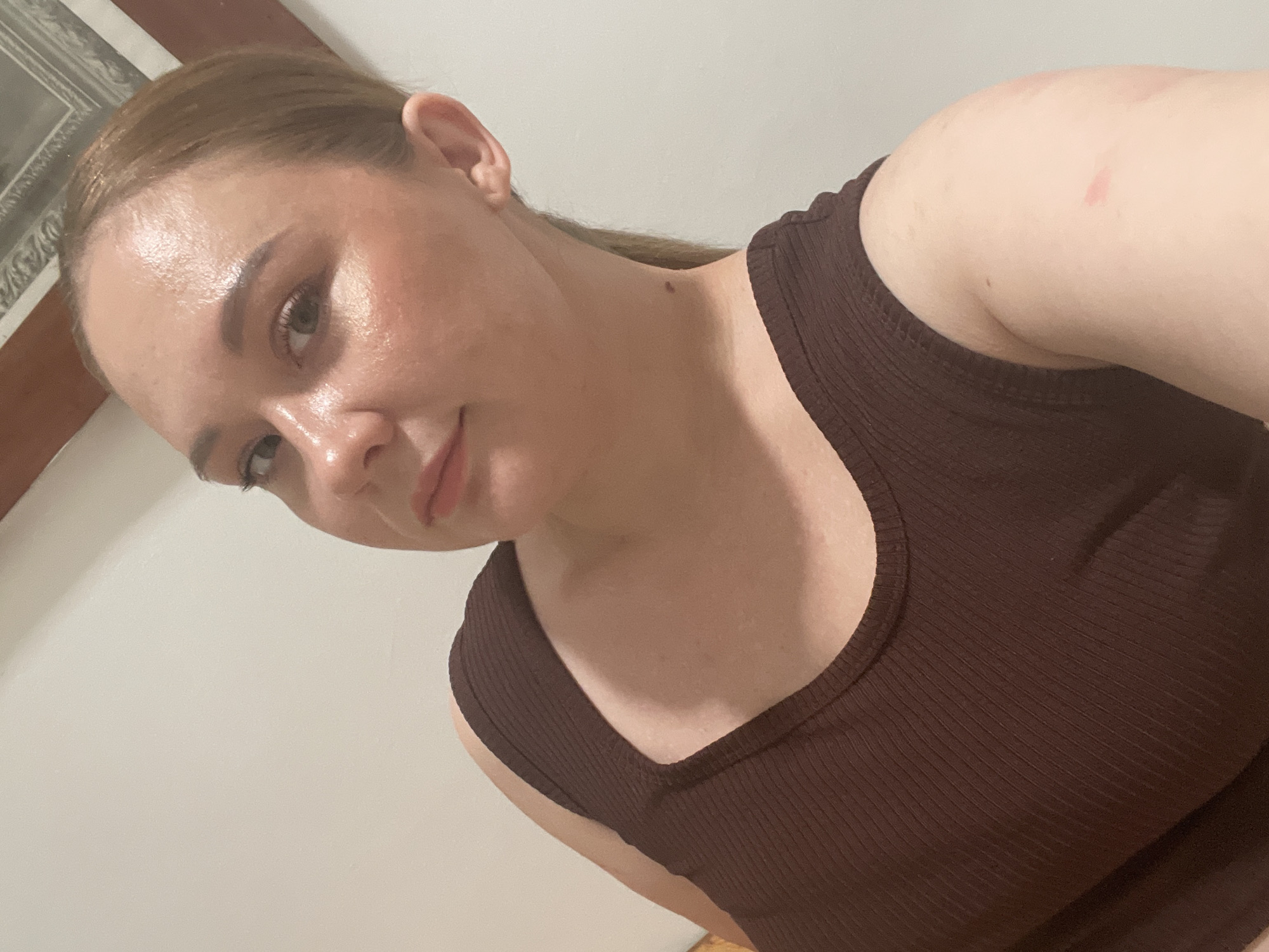 A woman with light skin and shoulder-length brown hair pulled back in a ponytail is shown in a close-up, from the chest up, against a plain off-white wall. She is wearing a brown ribbed tank top. The lighting in the image is warm and causes a sheen on her forehead and cheeks. She is looking slightly off-camera to her left. A faint blush is visible on her cheeks. A small dark mole is present on the side of her neck, near her ear. There are reddish marks on her right upper arm. The background shows a portion of a framed picture with a dark wood frame mounted on the wall. The scene is indoors.