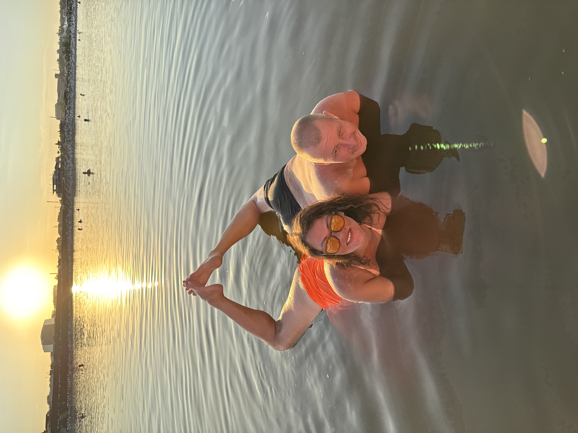 Two individuals, a woman and a man, are submerged in the calm, reflective waters of Lake Razval in Sol-Iletsk, Russia, during sunset. The woman, positioned to the left, wears an orange swimsuit and sunglasses, with her legs bent upwards, feet forming a V-shape. The man, to her right, wears black swim trunks. Both are lying on their fronts, partially submerged, facing the camera. Their reflections are visible in the water, creating a mirrored effect. In the background, the sun is setting, casting a golden glow on the water and the distant cityscape, which includes buildings and a structure resembling bleachers. Several other people are scattered in the lake, some appearing to swim or stand in the water. The overall atmosphere is serene, with the rippling water catching the warm light of the setting sun.