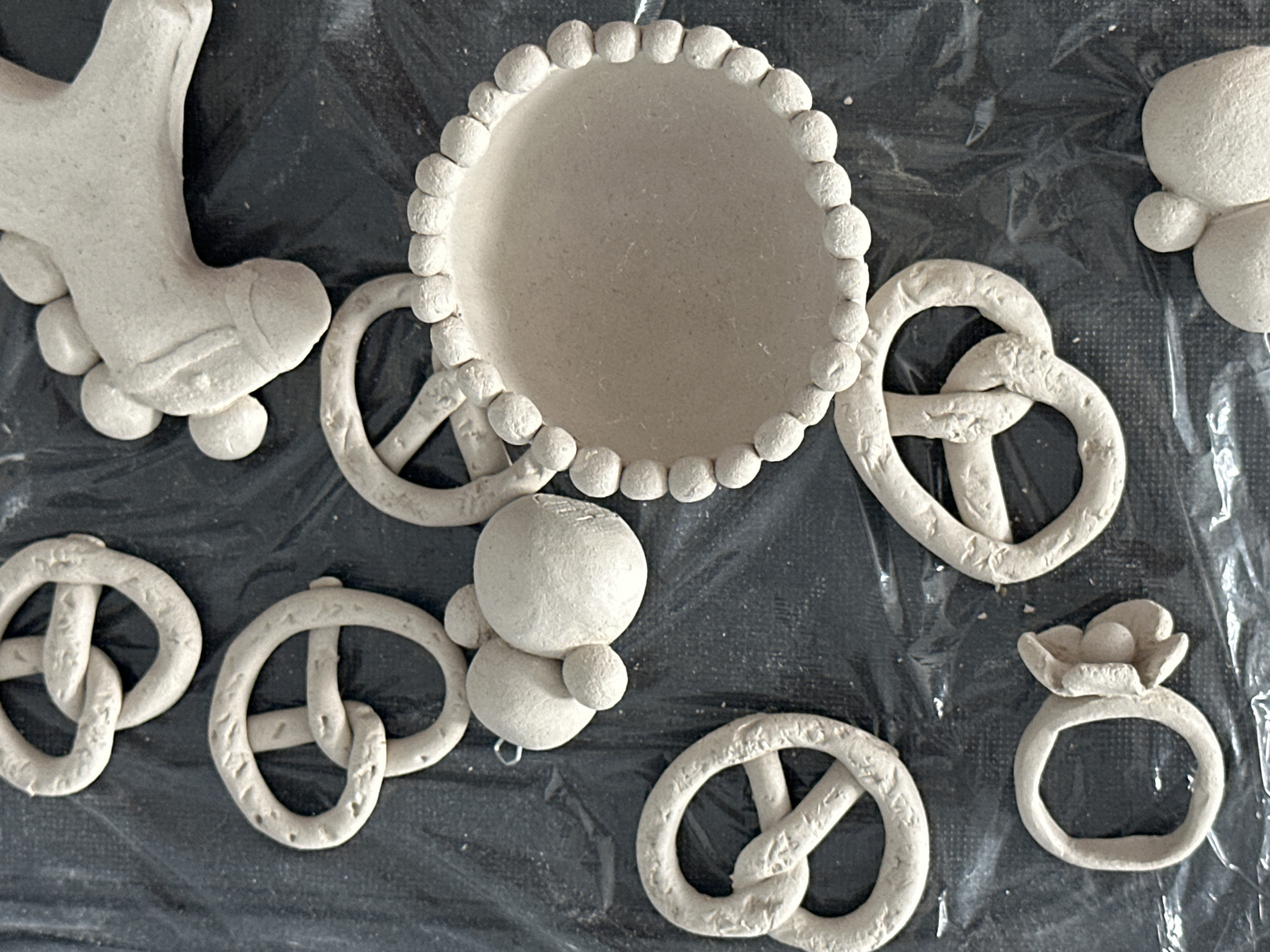 The image displays a collection of unbaked clay objects, predominantly pretzels, arranged on a black, crinkled surface, likely plastic sheeting. There are at least five pretzel shapes visible, varying in size and orientation. In addition to the pretzels, there is a small, circular dish with a beaded edge, a cluster of three spherical shapes that might represent grapes or berries, and a partial view of a clay horse's head. One pretzel has a small loop attached, suggesting it might be intended as an ornament. The lighting is overhead, casting subtle shadows that highlight the texture of the clay. The scene is static and appears to be a flat lay of craft items. There is no indication of Nevinnomyssk, Russia, or any specific location within a city.