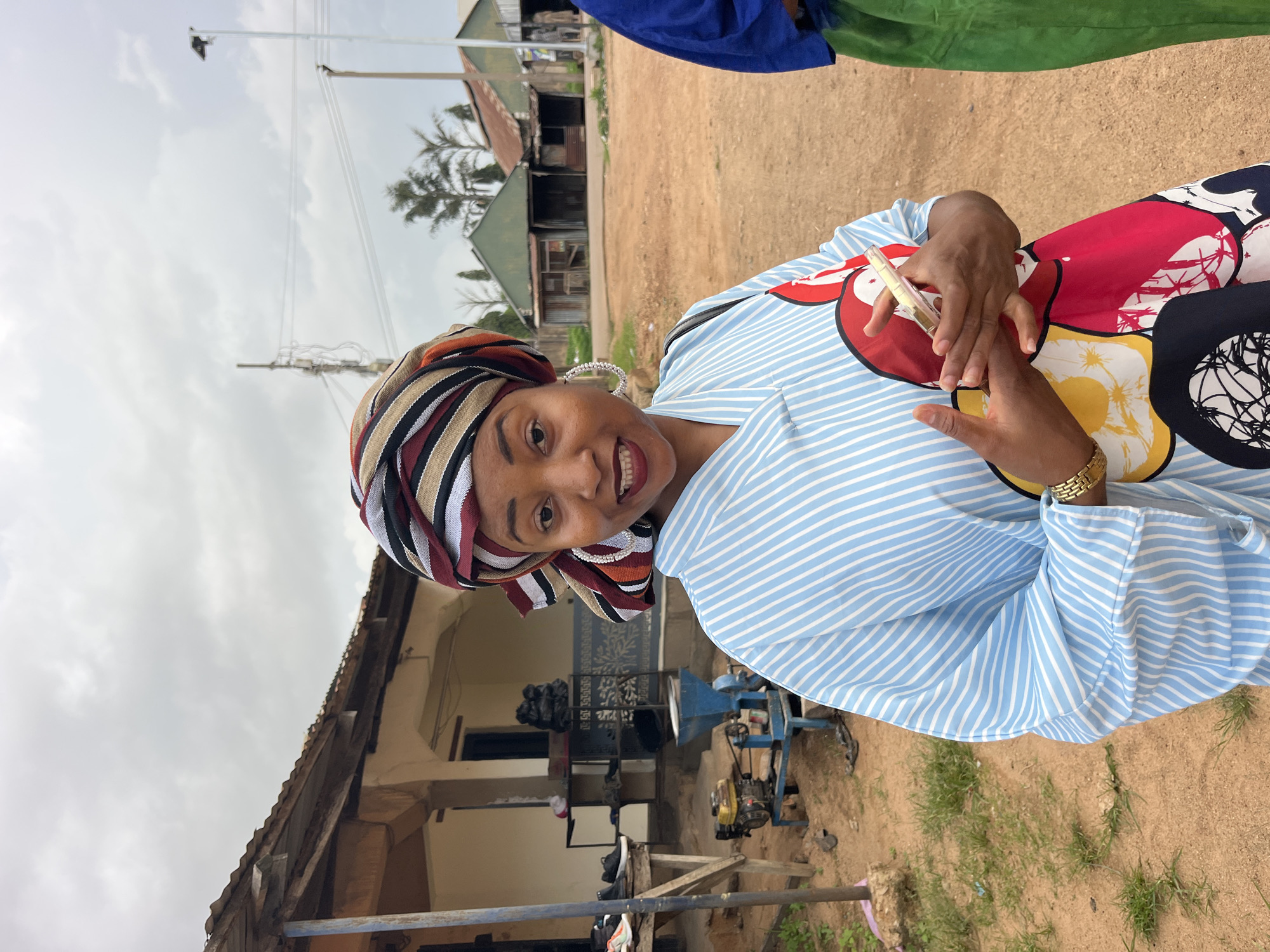 An African woman smiles at the camera, holding a clear object in her hands, as she stands in front of a building. She wears a blue and white striped abaya with a colorful headscarf. In the background, a yellow and blue machine sits on the ground, and in the distance, several small structures and trees are visible. The ground is a mixture of dirt and grass, and the sky is overcast. The scene appears to be an outdoor market or a rural village.