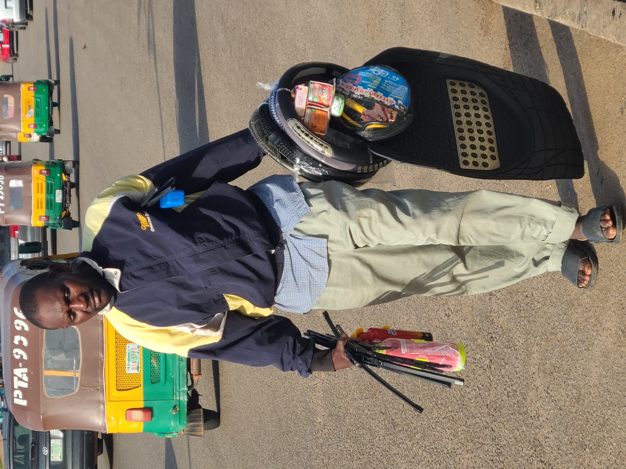 A man of African descent walks on a paved road in Jos, Nigeria, carrying items for sale. He is wearing a dark blue jacket with yellow and white accents, a light blue checkered shirt, and tan pants. On his right arm, he carries a helmet containing small cans and other packaged goods, along with a black rubber mat tucked underneath. In his left hand, he holds a bundle of what appear to be windshield wiper blades and a red fire extinguisher. Behind him, several colorful auto-rickshaws, commonly known as "keke napep," are visible on the road, suggesting a commercial area or busy street. The lighting indicates it is daytime, with shadows cast on the ground. The environment appears to be an urban setting with asphalt pavement.