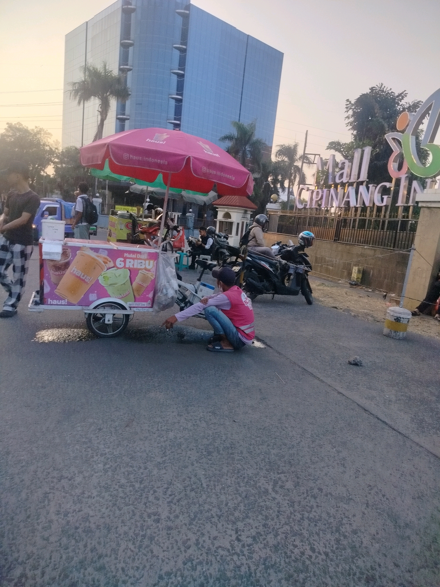 An exterior, ground-level, medium shot captures a street scene in front of Mall Cipinang Indah, East Jakarta, Indonesia. The late afternoon sun casts a warm glow on the scene. In the foreground, a person wearing a pink vest over a blue shirt and jeans squats down on the asphalt road, appearing to hold a cigarette and looking towards a beverage cart. The cart, adorned with colorful images of drinks and the "haus!" logo, is positioned on the right side of the frame. Behind the cart, several motorcycles are parked, and a man in a black shirt and checkered pants walks away from the viewer on the left. In the background, a tall, modern glass-faced building rises above palm trees and other foliage. A large, illuminated sign for "Mall Cipinang Indah" is prominently displayed. The road surface is textured with an irregular pattern, and a white cylindrical object with yellow bands stands near the right edge of the frame.