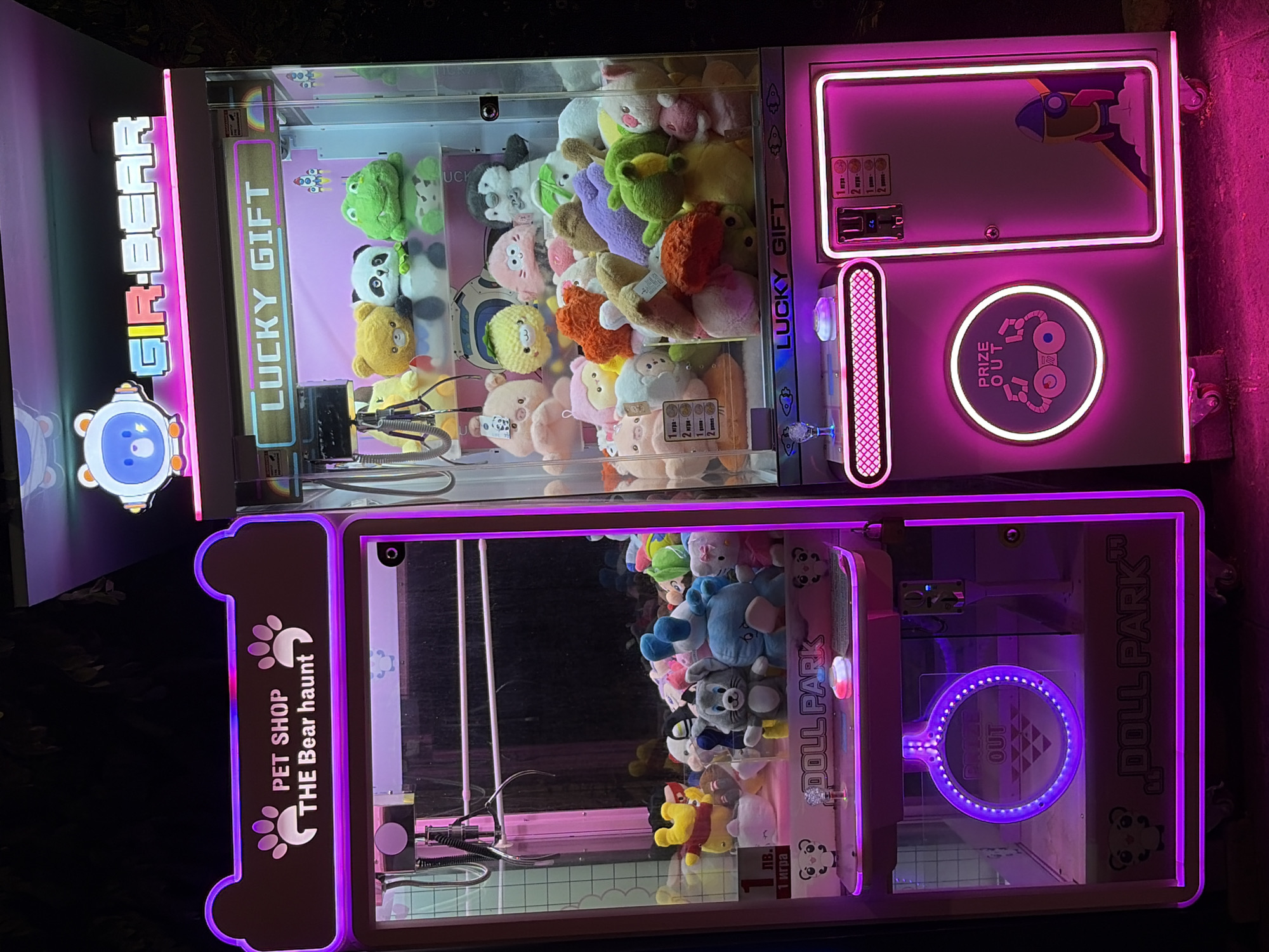 The image shows two brightly lit claw machines at night, likely outdoors, with neon pink and purple lights illuminating the scene. The machine on the left is labeled "PET SHOP THE Bear haunt" and "DOLL PARK". It is filled with various stuffed animals, including Winnie the Pooh, a grey rabbit, and a blue bear. The machine on the right is labeled "GIR-BEAR" and "LUCKY GIFT". It also contains a variety of plush toys, such as pandas, frogs, and bears. The machines appear to be in working condition, with a crane mechanism visible inside the left machine and a prize dispensing area on the right. The surrounding environment is dark, suggesting it is nighttime, and some foliage is visible in the background. The machines are on wheels, indicating they are movable. This scene is located in Obzor, Bulgaria, a coastal town known for its tourist attractions and amusement facilities.
