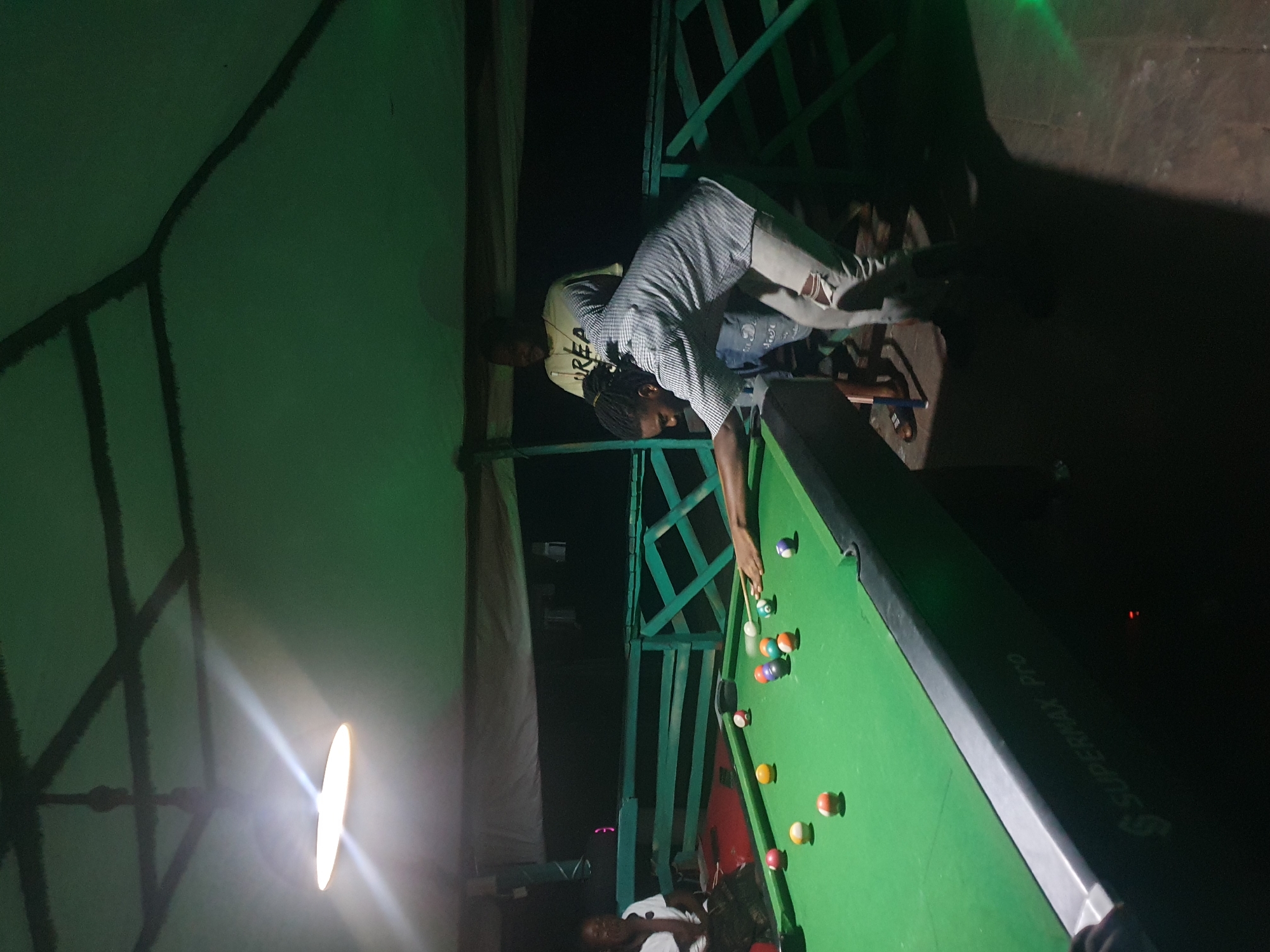 The media file shows a dimly lit outdoor scene at night, likely in Lokoja, Nigeria. The primary subjects are two individuals playing a game of billiards on a green pool table. One person, wearing a striped shirt and ripped jeans, is bent over the table, cue in hand, actively engaged in taking a shot. Another person, wearing a yellow shirt with visible text, stands behind them, observing the game.
The setting appears to be an informal gathering area, possibly a backyard or a patio, with a green, angular structure in the background. A bright, circular light source, possibly a lamp, is also visible on the left side of the frame, casting a strong glare. The ground is a dark, textured surface. The overall mood is relaxed and recreational.
A prominent object in the scene is the pool table, with several balls scattered across its surface. The balls are a mix of solids and stripes, with colors like red, yellow, orange, and green visible. The pool cue is a long, thin stick. On the side of the pool table, the text "SUPERMAX PRO" is visible, indicating the brand of the table. There is no clear indication of weather conditions, but the nighttime setting suggests it is not actively raining. The focus is on the game and the interaction between the players.
