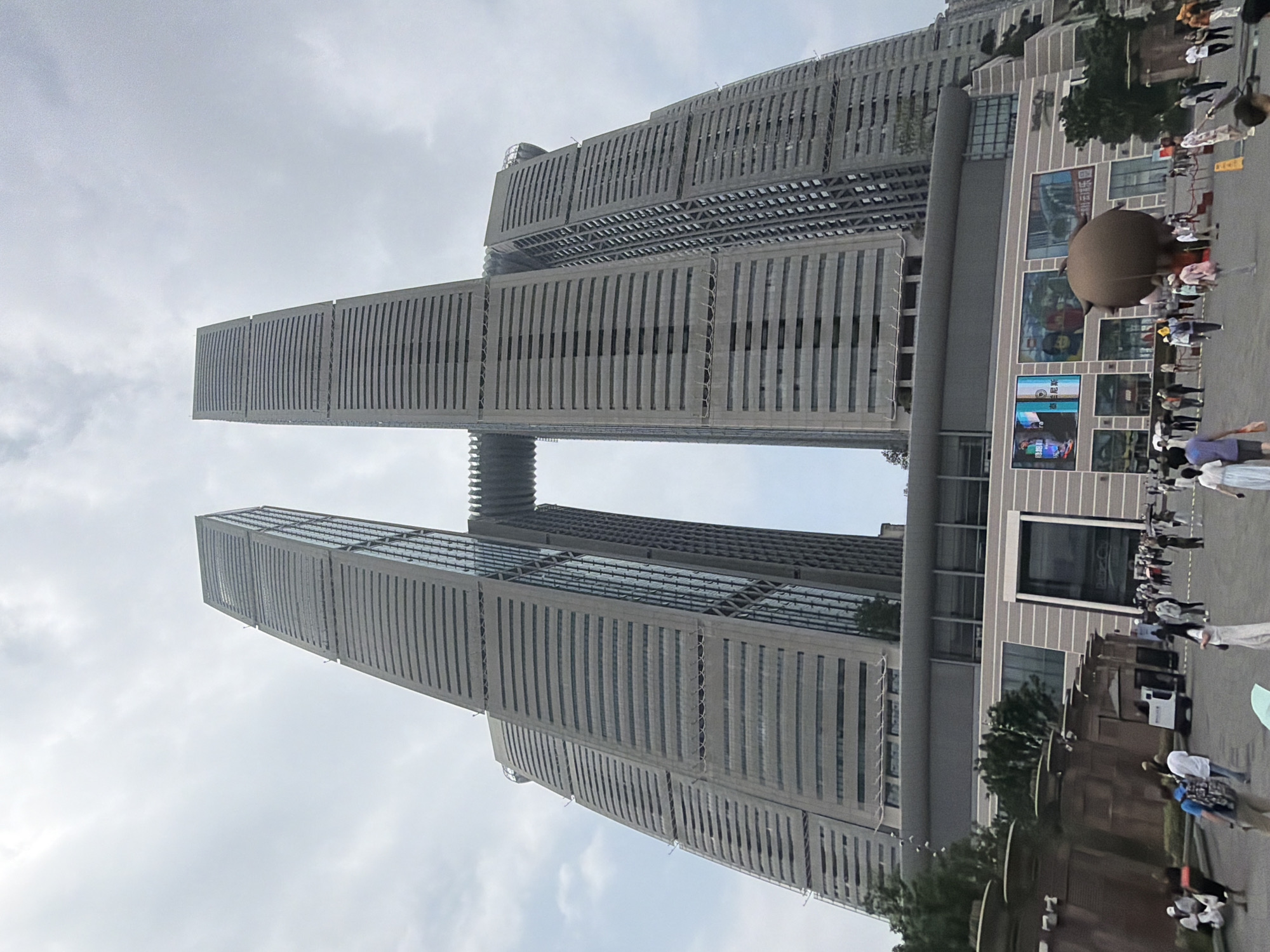 This is a wide-angle, low-angle shot of the Chongqing Chaotianmen Yangtze River Bridge, also known as the Raffles City complex, in the Yuzhong District of China. The complex is comprised of four interconnected skyscrapers, with a skybridge connecting two of the tallest towers. The buildings have a gray, segmented facade with horizontal window bands. The sky is overcast and a light gray. In the foreground, a plaza area with a brown, spherical inflatable sculpture is visible. People are walking around the plaza, some are sitting on benches, and there are some vehicles present, including a small white cart. The overall scene depicts a busy urban environment with modern architecture.