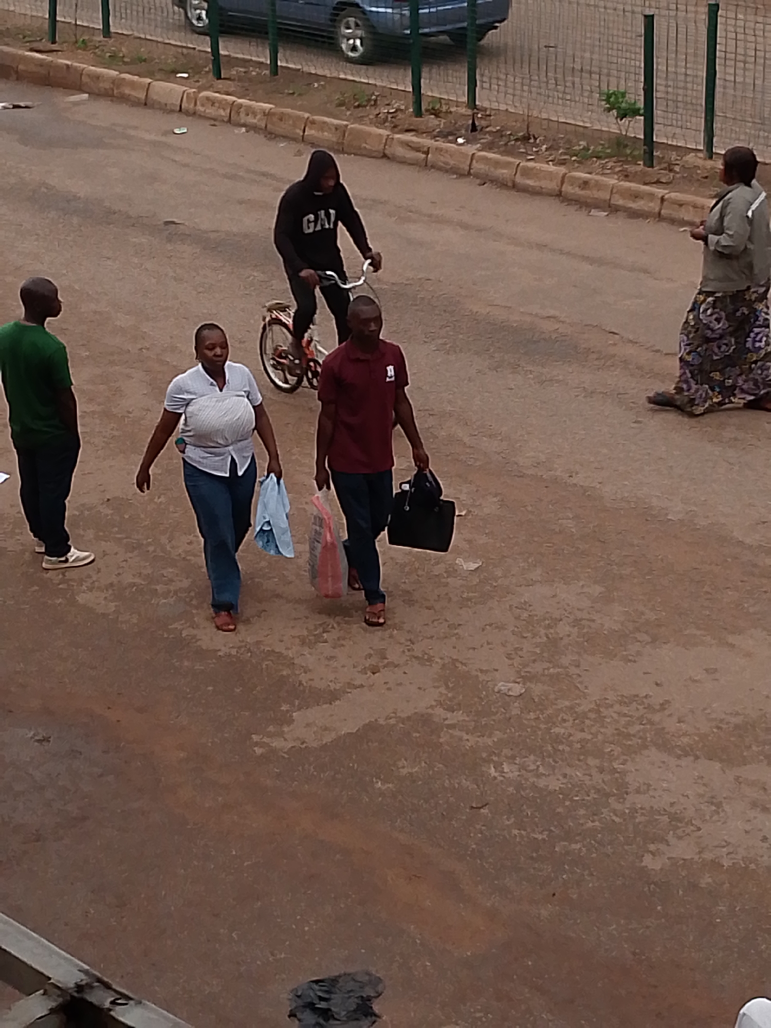 The image depicts an outdoor scene in Bukuru, Nigeria, featuring several individuals in a street environment. In the foreground, a man in a green t-shirt and dark trousers stands facing left. Beside him, to the right, a woman in a light-colored collared shirt and blue jeans walks forward, carrying a light blue cloth in her left hand and a plastic bag in her right. Another man, wearing a maroon t-shirt with a white emblem and dark trousers, walks beside her on the right, carrying a black bag. Further back, a person in a black hooded sweatshirt rides a bicycle, facing forward. On the far right side of the image, a woman in a light-colored jacket and a patterned skirt stands facing left. The ground is paved with asphalt, and a curb with rocks borders the street. A metal fence and some vegetation are visible in the background, along with a dark-colored vehicle. The overall scene suggests a casual, everyday moment in a Nigerian urban setting.