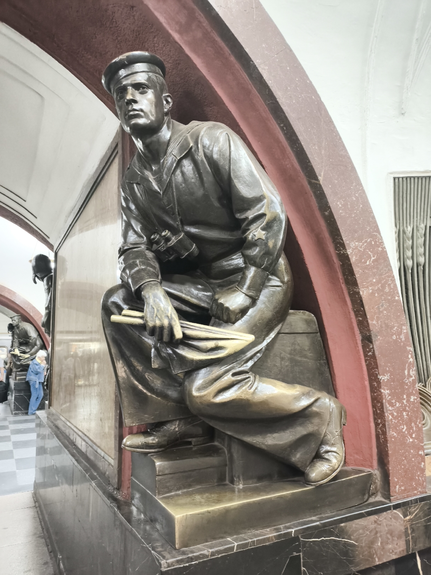 The image shows a close-up of a bronze statue of a man in a sailor's uniform. He is seated, with one leg bent and the other extended. He is wearing a sailor's cap, a loose-fitting jacket, and wide-legged trousers. His hands are resting on his lap, holding what appears to be a rolled-up document or flag. He is also wearing binoculars around his neck. The statue is positioned within an architectural setting featuring arched red and white structures, and a dark marble base. In the background, other figures and architectural elements are visible, including what appears to be a checkerboard tiled floor and another statue of a person. The lighting suggests an indoor environment. The statue appears to be part of a larger installation or memorial. There is no visible text in the image.