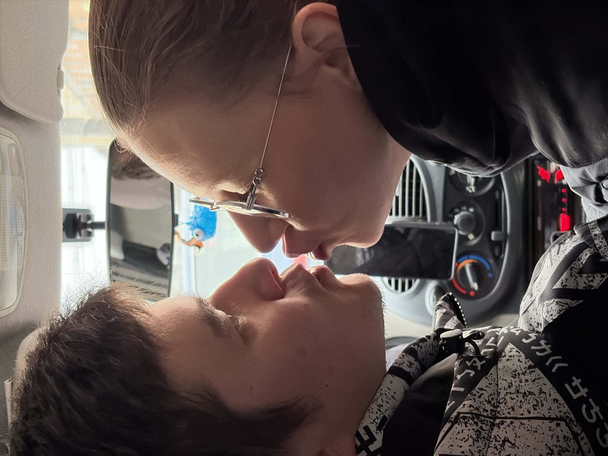 This intimate photograph depicts two individuals sharing a kiss or an embrace inside a vehicle, likely a car or van. The image is oriented sideways, suggesting the subjects are lying down or the photo was taken at an unusual angle. Bright natural daylight illuminates the scene, indicating it is daytime.
The person positioned above has light brown hair, wears distinctive thin metal-framed glasses, and a simple black top. Their face is angled downwards towards the other person. The second individual, positioned below, has darker hair and is looking up. They wear a black and white patterned garment, possibly a hoodie, adorned with geometric designs and stylized Japanese characters including "カ ワ 《 セ さ リ". A small black clip is also visible on their clothing.
In the background, elements of the car's interior are visible, including a dashboard with a climate control dial showing red and blue indicators, air vents, and a rearview mirror. A partial view of a ceiling light fixture is on the left. Through a window, a small blue bird-shaped ornament with an orange beak is discernable. The scene captures a tender and affectionate moment between the couple. No specific cues linking the location to Izhevsk, Russia are visible within the image itself.
