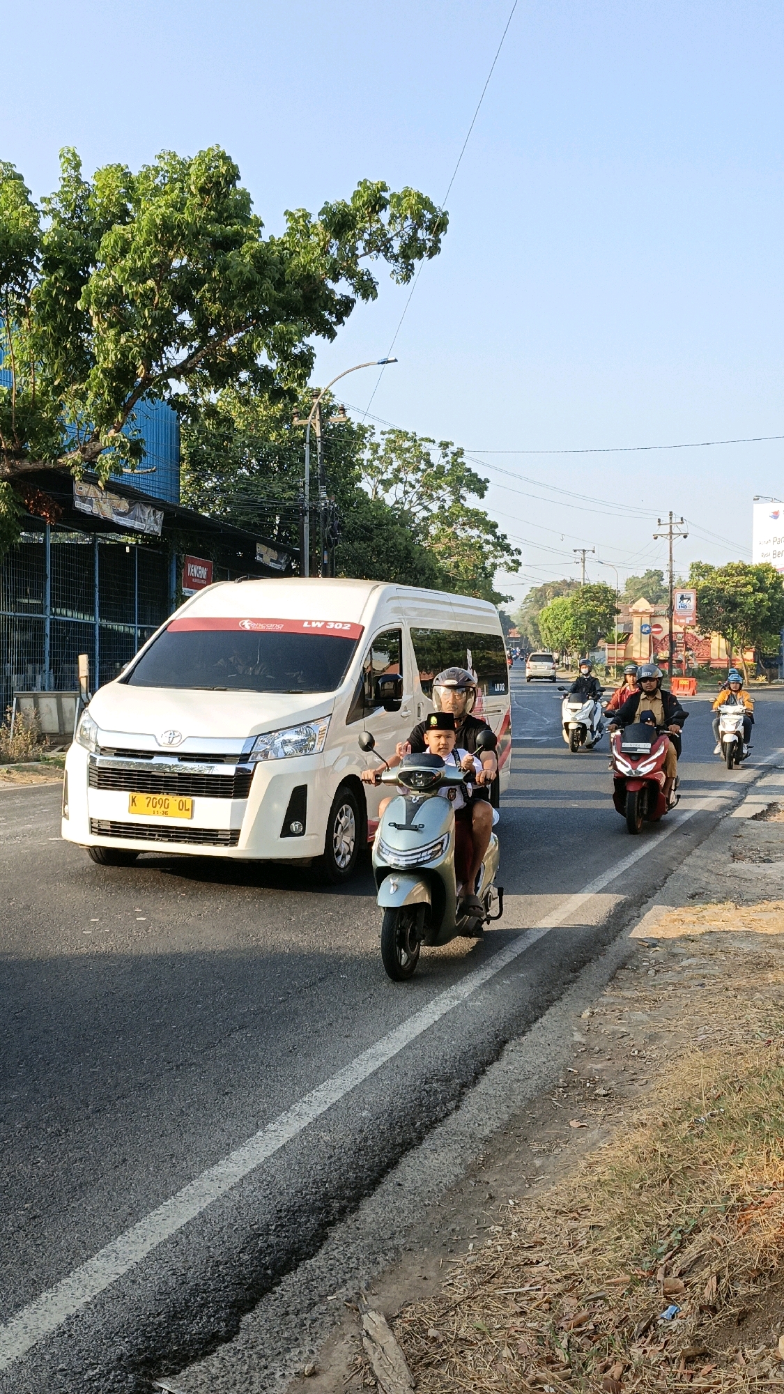 In Kuwasen, Indonesia, a white van with "LW 302" visible on its side is traveling on a paved road. A man and a child on a light-colored scooter are in the foreground, traveling in the same direction as the van. The child, wearing a dark hat and a white shirt, is in front, and the man is behind him. Several other motorcycles are also on the road, with riders wearing helmets. The environment appears to be a street with trees and buildings lining the side of the road. The sky is clear and blue.