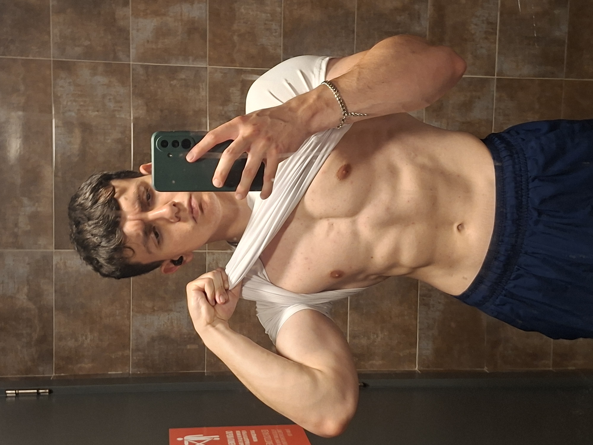 A man is posing for a selfie in what appears to be a public restroom. He is fair-skinned with dark hair and wearing dark blue athletic shorts. He is holding a green smartphone and lifting up his white t-shirt to reveal his abdomen. The background consists of brown square tiles, and a sign with red text is partially visible at the bottom of the frame. There is no clear indication of the location in Plovdiv, Bulgaria from this image.
