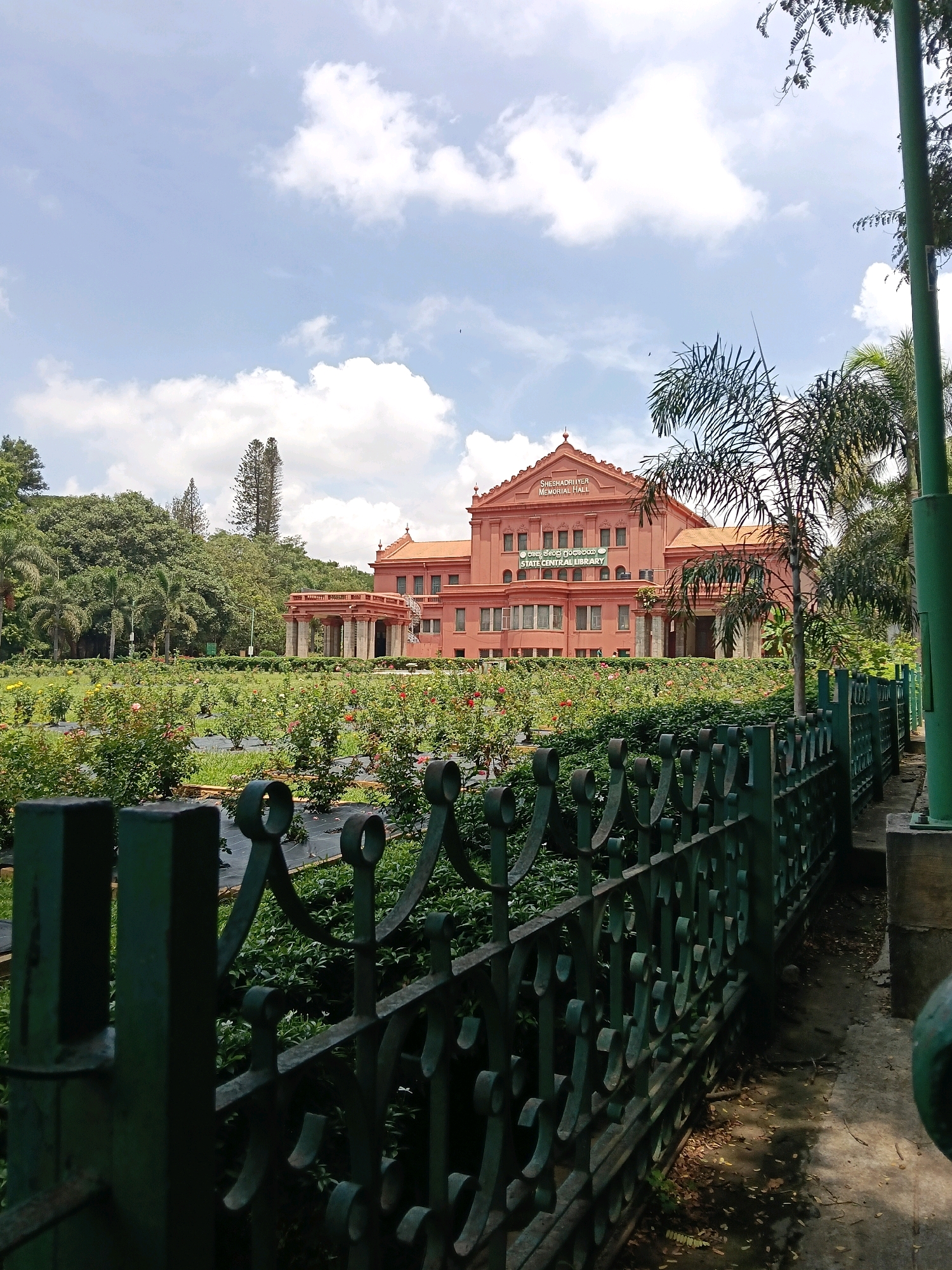 The image depicts the Seshadri Iyer Memorial Hall, a historical building in Cubbon Park, Bengaluru, India. The building, painted in a salmon pink hue, is visible in the background, featuring classical architecture with columns and a prominent pediment. In front of the hall lies a well-maintained rose garden, with rows of rose bushes in bloom, some covered with black netting. A dark green, ornate metal fence runs along the foreground, partially obscuring the garden and a concrete path. Lush green trees and palm trees surround the building and garden, with a bright, cloudy blue sky overhead. The scene appears to be a peaceful, daytime view of a public space.