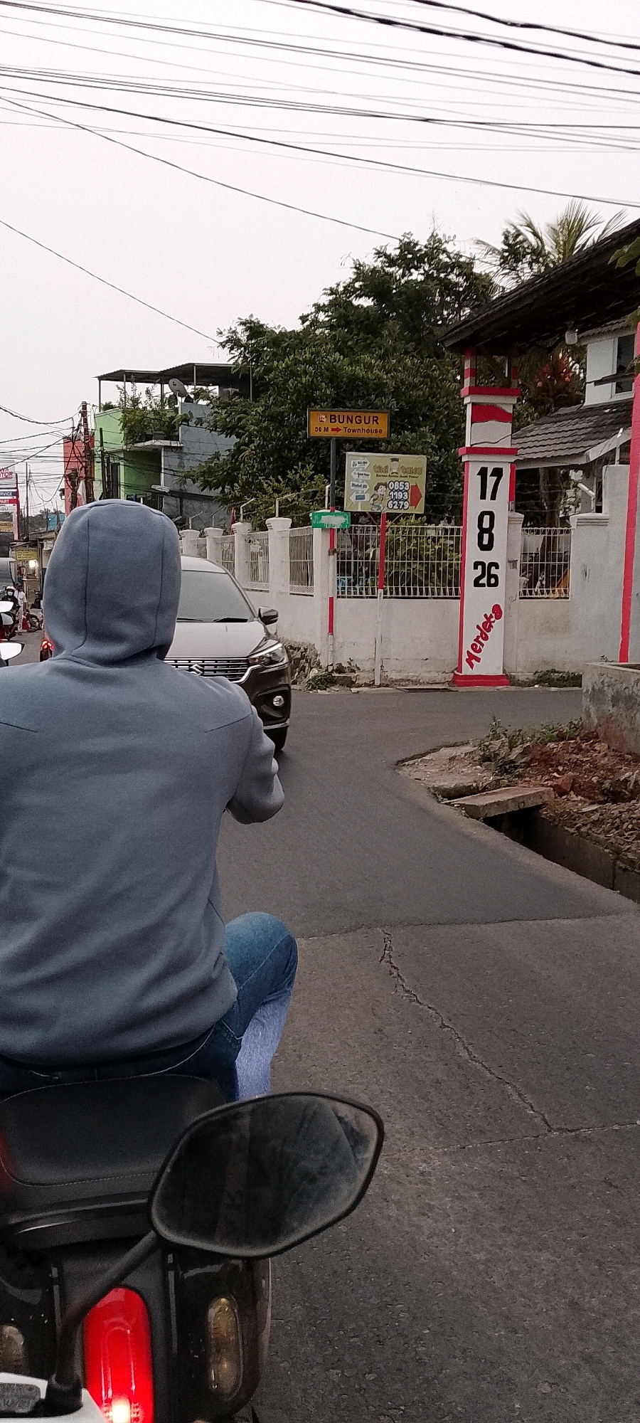The image shows a street scene in Depok, Indonesia, captured from the perspective of someone riding a motorcycle. In the foreground, the back of a person wearing a grey hooded sweatshirt and blue jeans is visible, seated on the motorcycle. The motorcycle's rear red taillight and a side mirror are also prominent.
The street itself is paved with asphalt, showing some cracks. To the right, a concrete drain runs alongside the road, with a small concrete channel. The road curves slightly to the right.
In the middle ground, a dark-colored car is driving on the road. Beyond the car, there is a white fence with decorative red elements. A white and red striped pillar stands at the entrance to a property, bearing the numbers "17", "8", and "26" stacked vertically, with the word "Merdeka" written in red on its side. Adjacent to the pillar is a building with a dark roof.
Further in the background, a yellow sign with red text reads "BUNGUR 50 M". Below this sign, another sign with multiple smaller signs attached features telephone numbers: "0853", "1193", and "6279". Trees and buildings are visible behind the fence. Overhead, numerous electrical wires crisscross the sky, indicating an urban or suburban environment. The sky is overcast, suggesting a cloudy day.