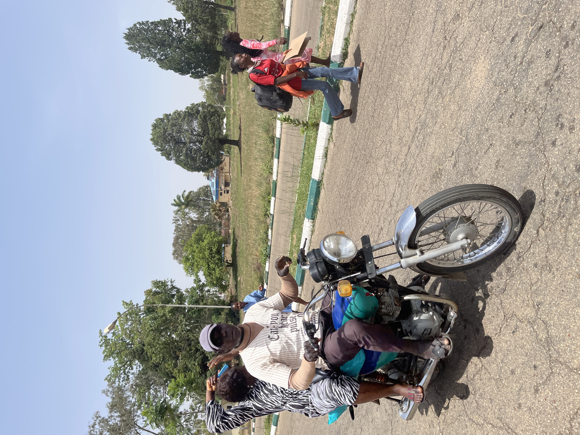 Two adults are on a motorcycle on a paved road. One adult is operating the motorcycle, wearing a striped shirt, grey pants, black gloves, a baseball cap, and sunglasses. The other adult, wearing a zebra-striped top, sits behind the motorcycle operator. In the background, two other adults are walking on the side of the road. The location appears to be the city of Jos, Nigeria, based on the environment and attire.
