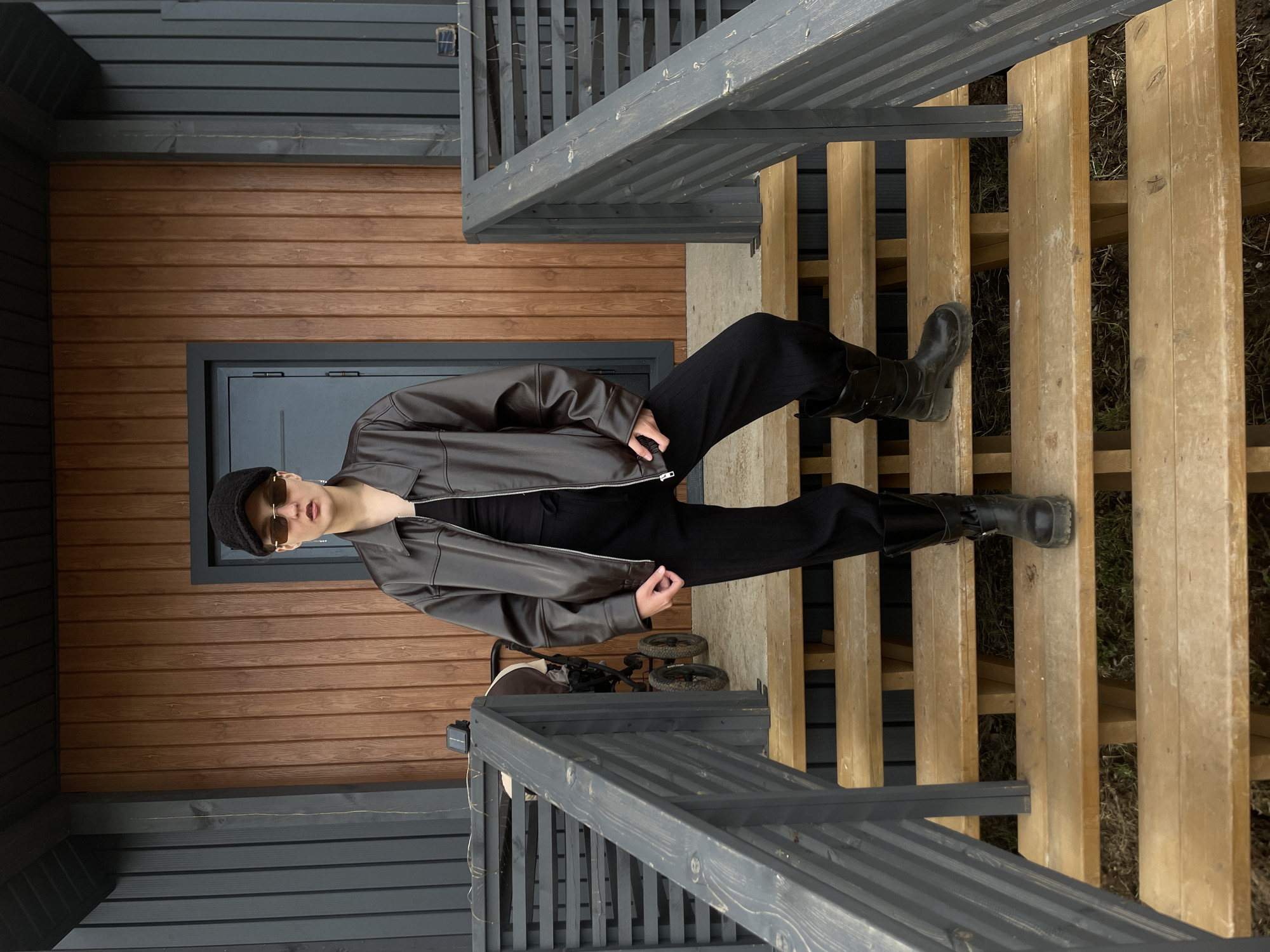 The image depicts a young person fashionably dressed, posing outdoors on a multi-level wooden structure in Подшивалово, Russia. The person, positioned centrally, wears a dark brown faux-leather jacket, black loose-fitting trousers, chunky black boots, a black flat-top cap, and rectangular sunglasses with light brown frames. They look directly at the camera with a composed, neutral expression, with one hand near their jacket's zipper.
The setting appears to be a porch or deck with descending wooden steps made of light-colored planks, flanked by dark gray painted wooden railings. Behind the person, to their left, is the exterior of a building featuring warm reddish-brown horizontal wooden siding topped by dark gray horizontal siding. A dark gray door frame is visible within this wall. To the right of the person, partially obscured by the railing and steps, is a small, two-wheeled cart. A small black solar panel is mounted on the right-hand railing. The ground visible between the steps is dry earth with sparse grass. The scene is well-lit, indicating daytime, likely under overcast skies, given the soft, even lighting. No discernible text is visible in the image.