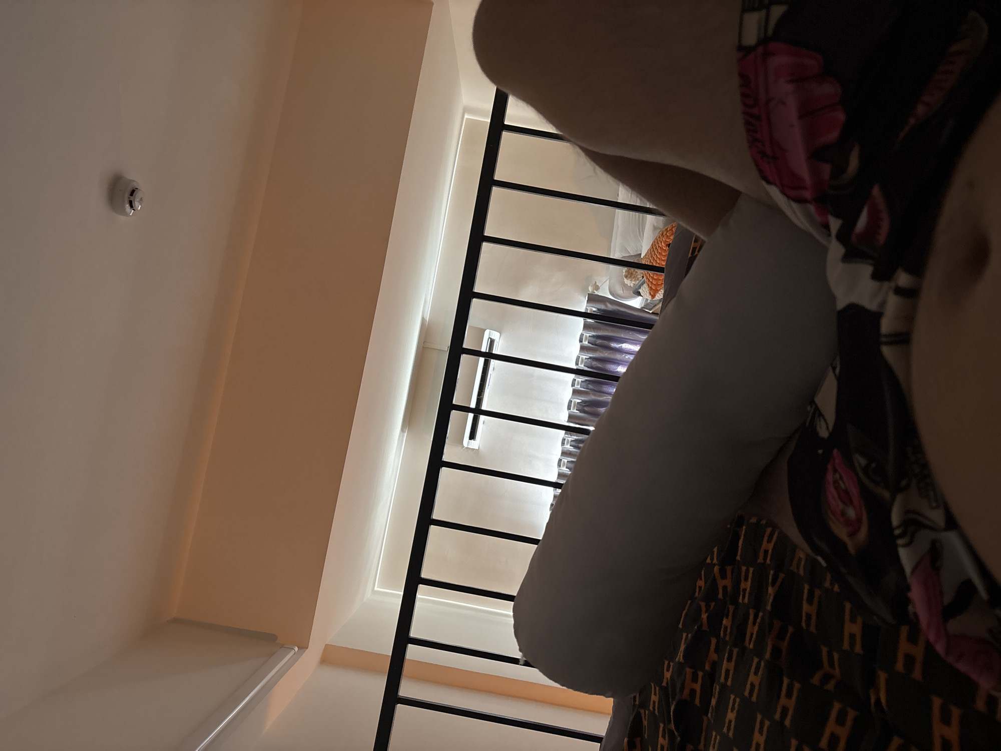 The image shows a partial view of a room with a white wall and a staircase with black railings. A person wearing patterned clothing is visible in the foreground. A smoke detector is mounted on the ceiling. The location is not specified.