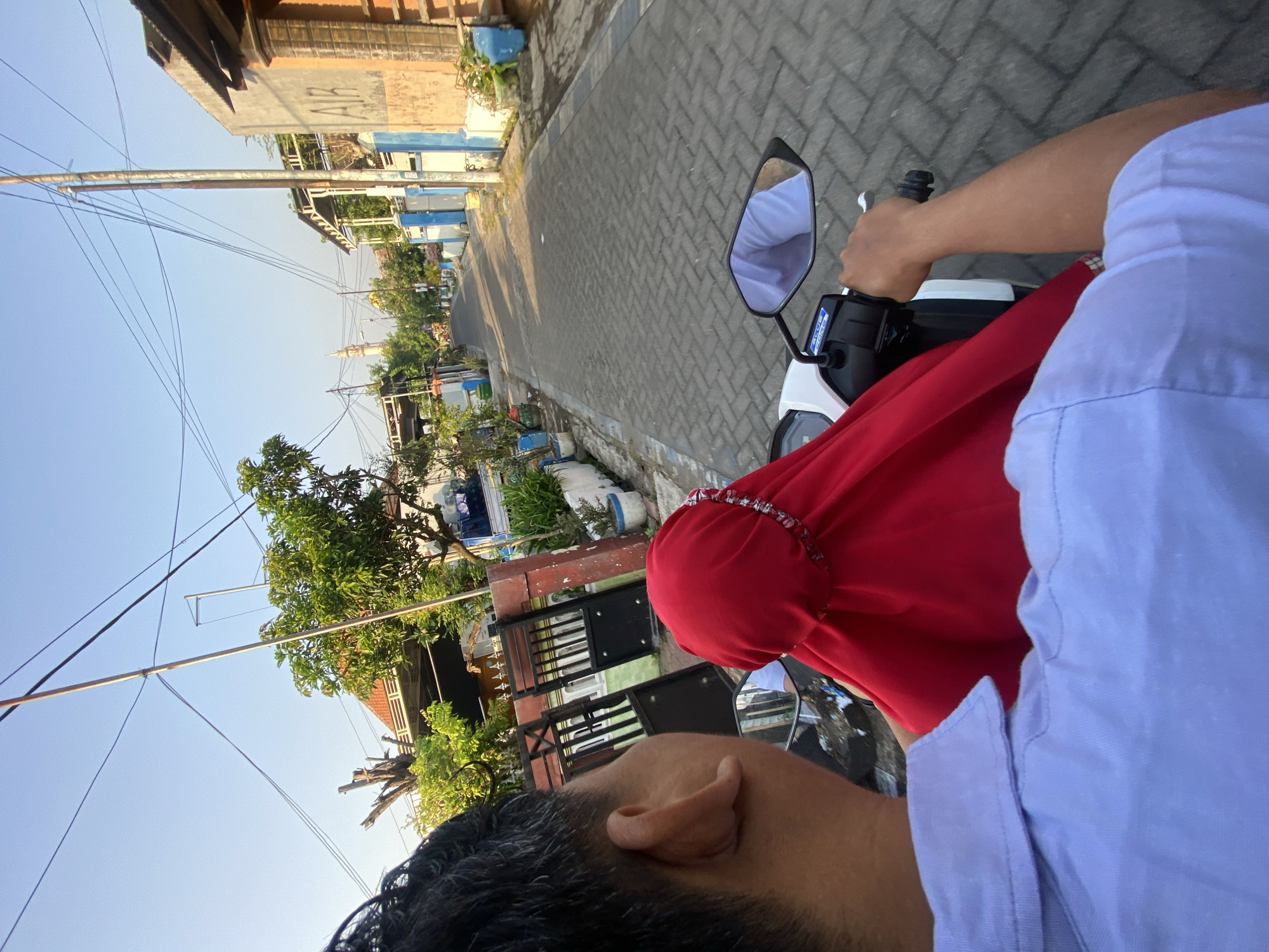 In Ngampelsari, Indonesia, two individuals ride a white scooter down a narrow, paved street lined with houses. The person in front, wearing a light purple collared shirt, is seen from the back, with their right hand on the scooter's handlebars. The passenger behind them is wearing a bright red hijab. The street is illuminated by sunlight, casting long shadows. Houses with various architectural styles and colors, some with metal gates and potted plants, flank the road. Trees and electrical wires are visible in the background, and a mosque's minaret can be seen in the distance. The overall scene depicts a typical day in a residential area of Ngampelsari.