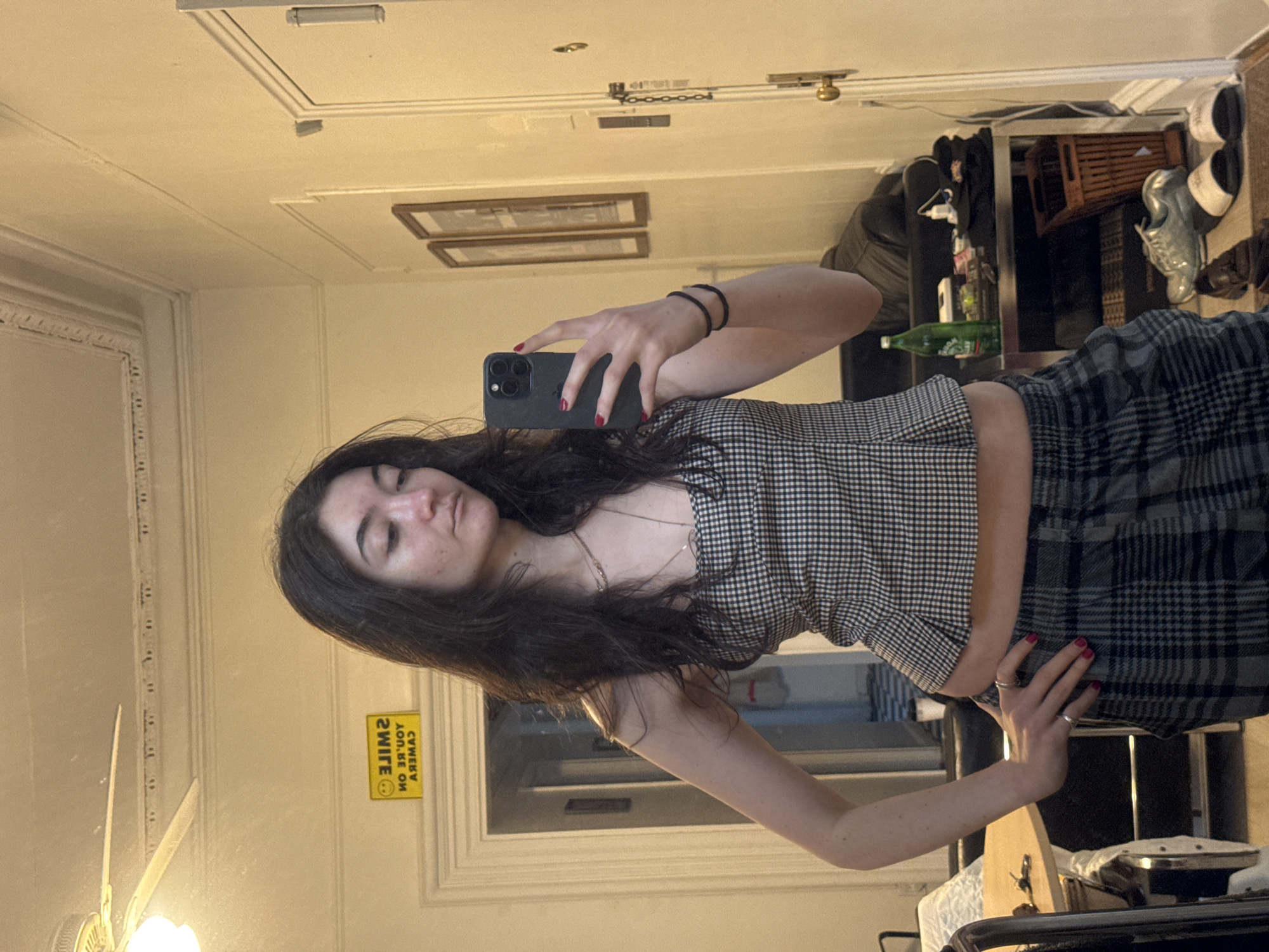 A woman is taking a selfie in a mirror. She is wearing a plaid top and skirt, and has dark hair. The mirror reflects part of a room with a window, and what appears to be a storage area with various items including shoes, a green bottle, and some clothing. A "smile, you're on camera" sign is visible in the reflection. The location is not discernible as being in New York City.