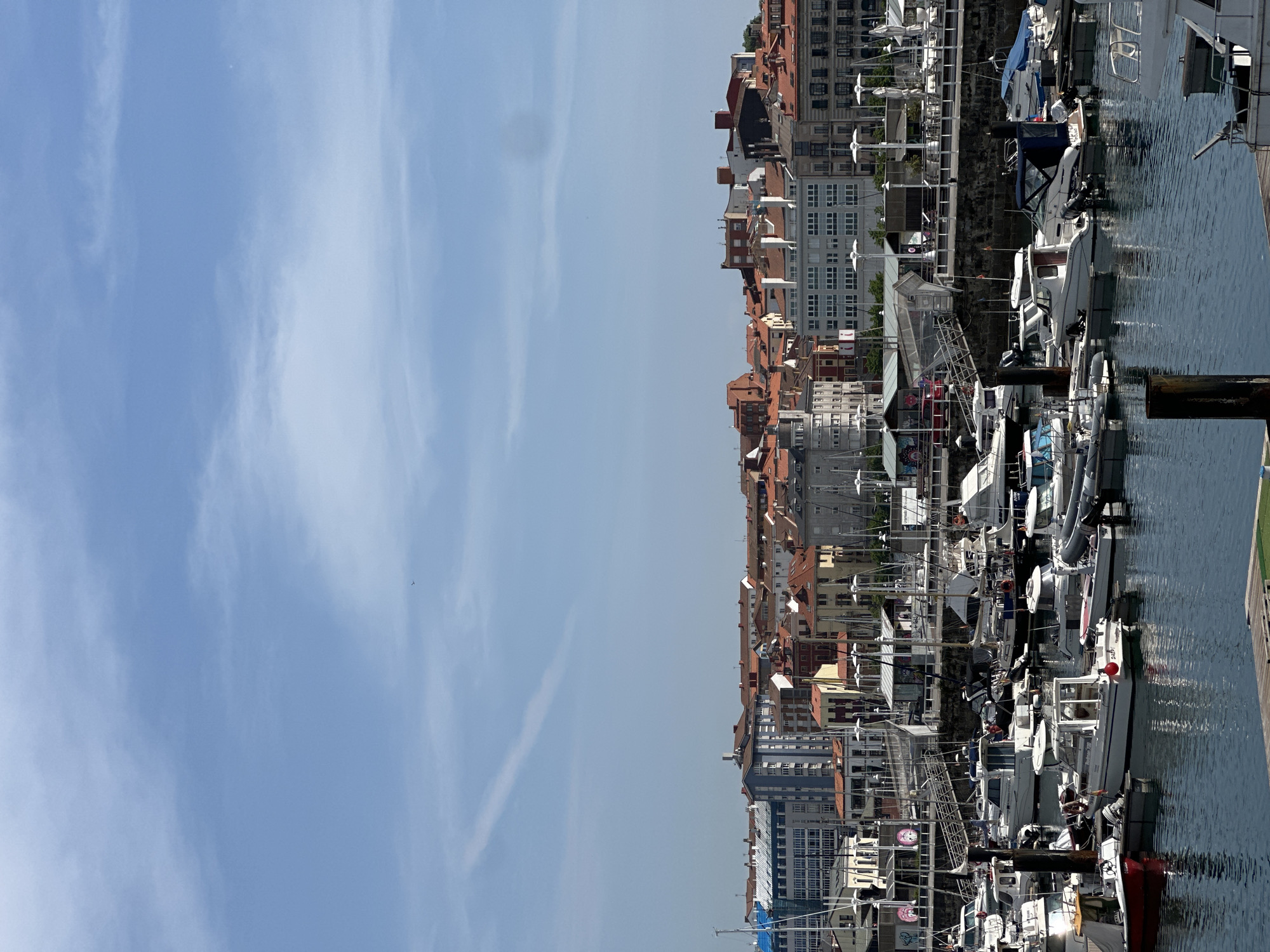 The image shows a marina filled with boats alongside a harbor in Gijón/Xixón, Spain. The cityscape includes buildings with orange tile roofs and white or grey facades. A clear blue sky with faint contrails dominates the left portion of the image. The water reflects the buildings and boats.