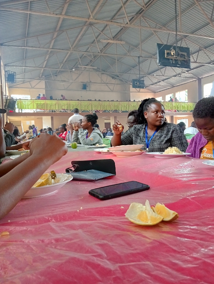 The image depicts a bustling communal meal taking place within a large indoor hall, likely in Riara Ridge, Kenya. The hall features a high, industrial-style ceiling with exposed metal trusses, from which large speakers or lights are suspended. The space includes a main floor and an upper balcony or mezzanine level, both occupied by numerous people seated at tables.
In the immediate foreground, a table covered with a crinkled, translucent red plastic tablecloth is visible. Several individuals are seated around this table, engaged in eating. On the left, the arms of a person are seen reaching for food on a white plate that contains what appears to be a meal with grains and slices of fruit, possibly lemons or limes. A black smartphone and a black wallet or clutch are resting on the red tablecloth. Further to the right, another black smartphone lies on the table, alongside two peeled orange segments.
In the midground, a woman with braided hair, wearing a patterned dark shirt and a blue lanyard, is seated at the table, looking attentively to her right while holding a piece of food. To her right, another person is partially visible, wearing a purple top with the word "Queen" faintly discernable on it. Throughout the rest of the hall, numerous individuals are seated at other tables on the main floor and along the upper balcony, indicating a large gathering. The hall appears to be illuminated by natural daylight entering from unseen windows. The scene suggests a busy event, possibly a conference, workshop, or community feast, where attendees are sharing a meal.