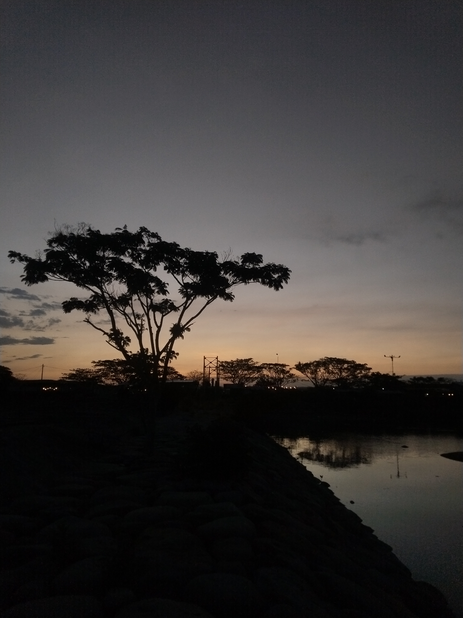 The image depicts a twilight scene in Lhokseumawe, Indonesia. A large tree is silhouetted against a sky transitioning from orange near the horizon to grey at the top of the frame. Below the horizon, other trees and distant lights are visible. Water, likely a river or pond, reflects the light and shapes. In the foreground is a dark, rocky embankment. No people are visible.