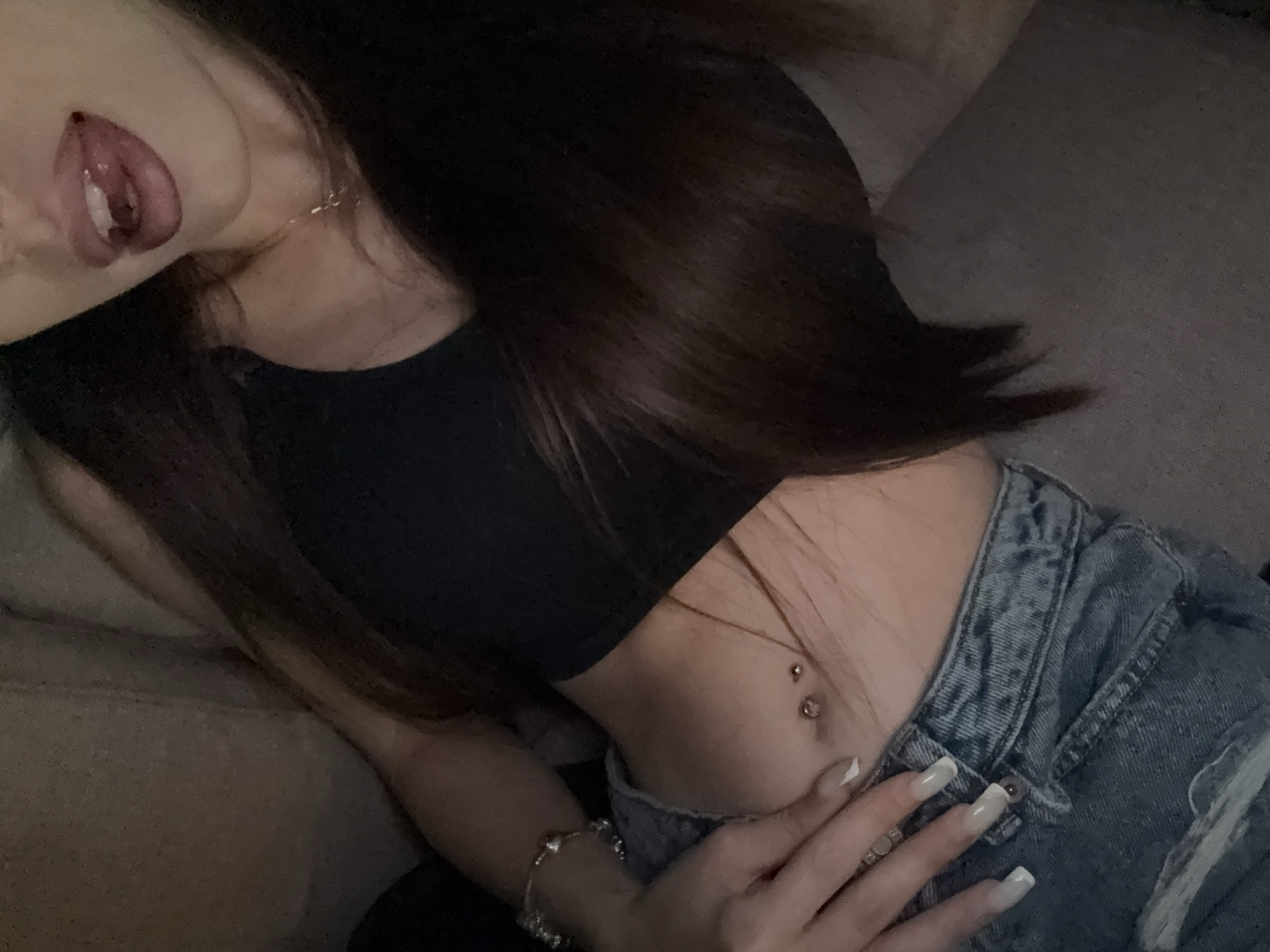 A close-up, overhead shot shows a person's midsection and mouth. The individual is wearing a black crop top, blue distressed denim jeans, and a silver bracelet on their left wrist. Their nails are long and painted with a light grey polish and white tips. The person has a belly button piercing with two pieces of jewelry. Their lips are slightly parted, with the tongue visible. The background appears to be a couch. The scene is set indoors in an unknown location.