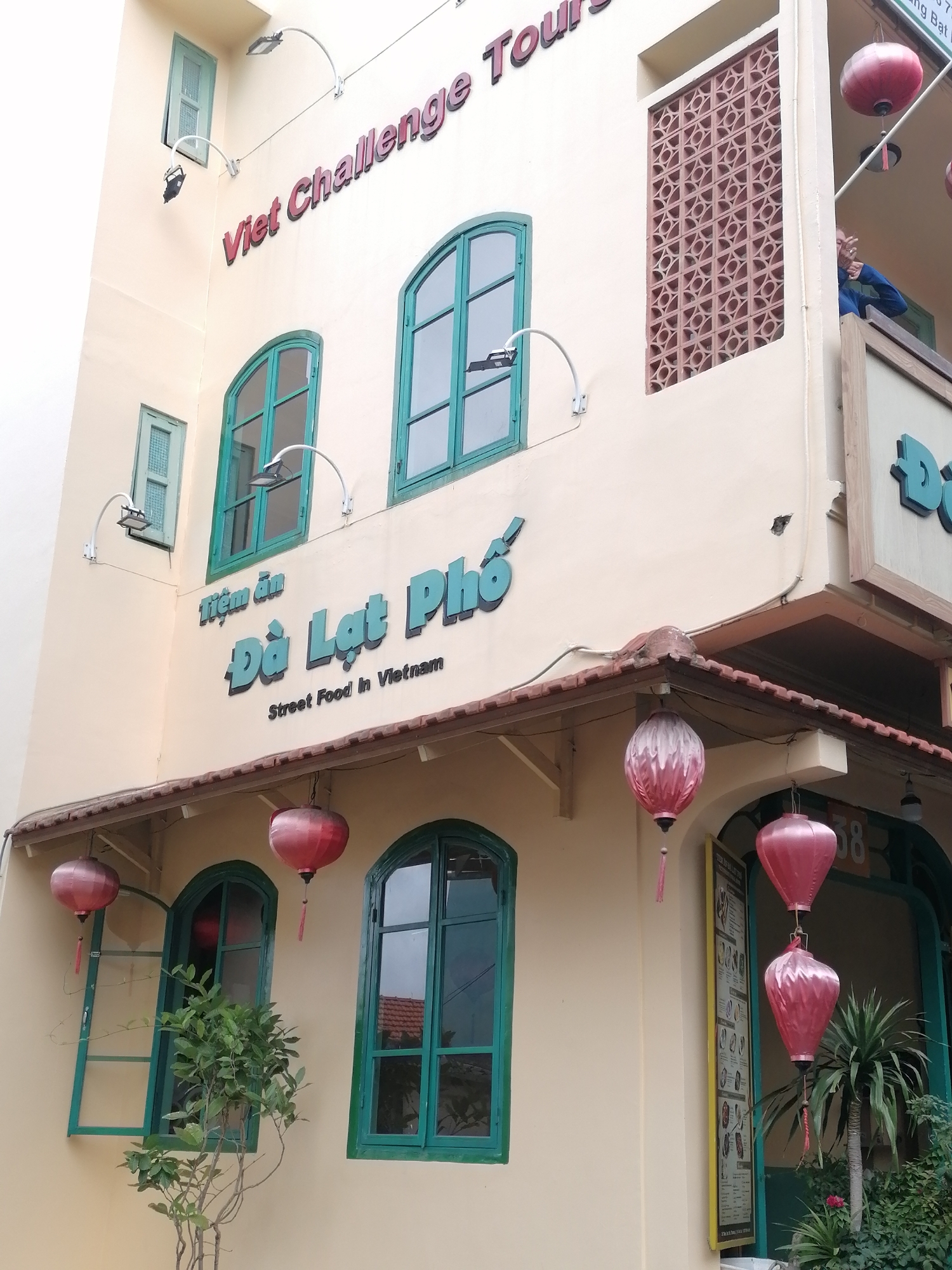 The image shows the exterior of a building in Da Lat, Vietnam, with a sign that reads "Viet Challenge Tours" and "Tiệm ăn Đà Lạt Phố Street Food in Vietnam." The building has light yellow walls and several windows with green frames. Some windows are open, and one has a small plant inside. Red and pink lanterns hang from the building's facade, and there is some greenery around the entrance. The overall impression is that of a street food establishment in a Vietnamese city.