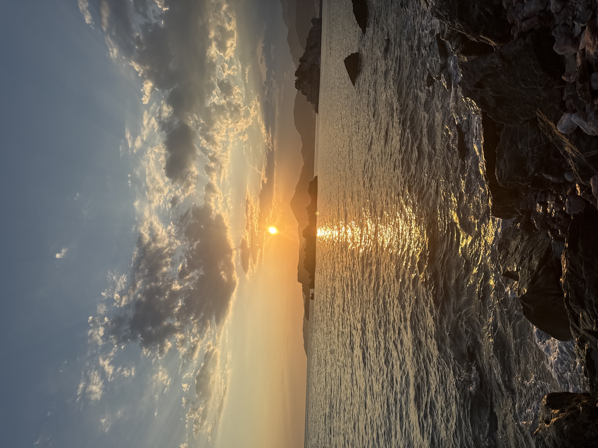 The media file depicts a sunset over the Adriatic Sea near Sveti Stefan, Montenegro. The scene is divided between the sky and the water. The sky is a canvas of soft blues and warm yellows, with scattered clouds catching the golden rays of the setting sun. These rays dramatically pierce through the clouds, creating a luminous effect. In the distance, silhouetted mountains form the backdrop, with a hint of buildings visible on one of them, suggesting a coastal town or village.
The sea occupies the right half of the frame, with its surface shimmering and reflecting the sun's glow, creating a dazzling path of light across the water. The texture of the water is rough, with many ripples indicating a gentle movement. To the far right and along the edge of the water, dark, rugged rocks are visible, some partially submerged. These rocks add a sense of natural texture and contrast to the smooth expanse of the sea and the soft colors of the sky.
The time of day is clearly sunset, given the low angle of the sun and the warm, diffused light. The weather appears calm, with the gentle ripples on the water suggesting a mild breeze. The location cues, such as the distinctive silhouette of the mountains and the coastal setting, combined with the provided context of Sveti Stefan, Montenegro, firmly place the scene in this picturesque European location. The overall mood conveyed is one of tranquility and natural beauty, with the sunset casting a serene and awe-inspiring atmosphere. There is no visible text in the media file.