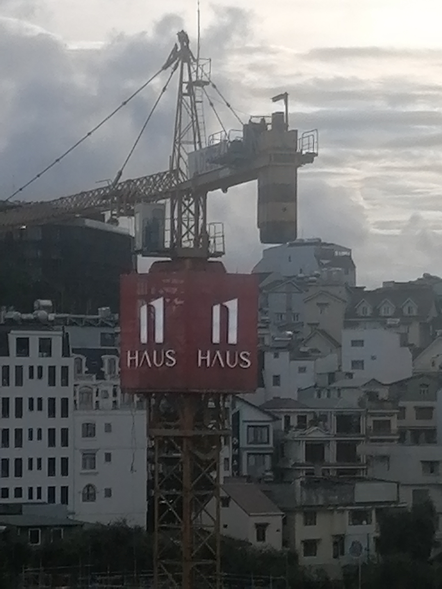 A construction crane is prominently displayed in the foreground of the image, with its yellow boom extending across the upper left portion. A red sign is attached to the crane's tower, featuring white numerals "11" and the text "HAUS HAUS" in white letters below. The background is filled with a dense cluster of buildings of varying sizes and architectural styles, indicative of a city skyline. Some buildings are white and modern with numerous windows, while others appear older, with pitched roofs and a mix of colors. The sky is overcast and cloudy, suggesting a dull or potentially rainy day. The overall scene captures an urban landscape under construction. Based on visual cues like the architecture and the presence of a construction crane, this scene is likely located in Da Lat, Vietnam.