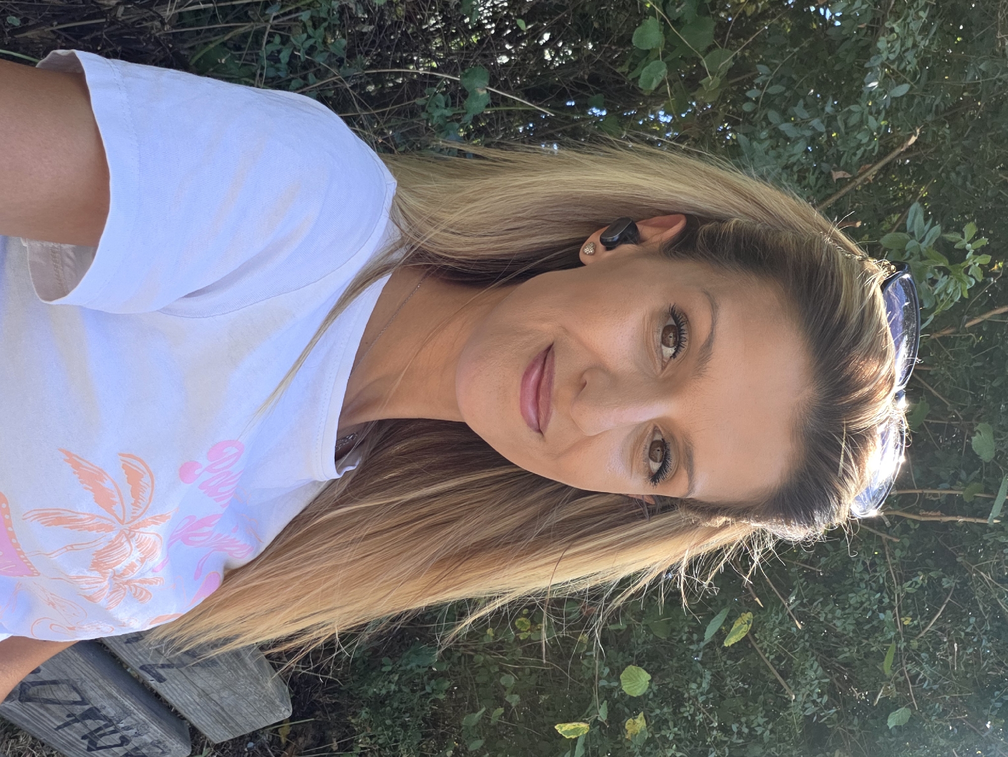A woman is posing for a selfie outdoors, seated on a wooden bench. She has blonde hair, is wearing a white t-shirt with a pink graphic of palm trees, and has sunglasses perched on her head. She is also wearing small black earbuds. The background consists of dense green foliage and trees, suggesting a park or natural setting within Munich, Germany. The lighting indicates it is daytime, with the sun casting highlights on her hair and the foliage.