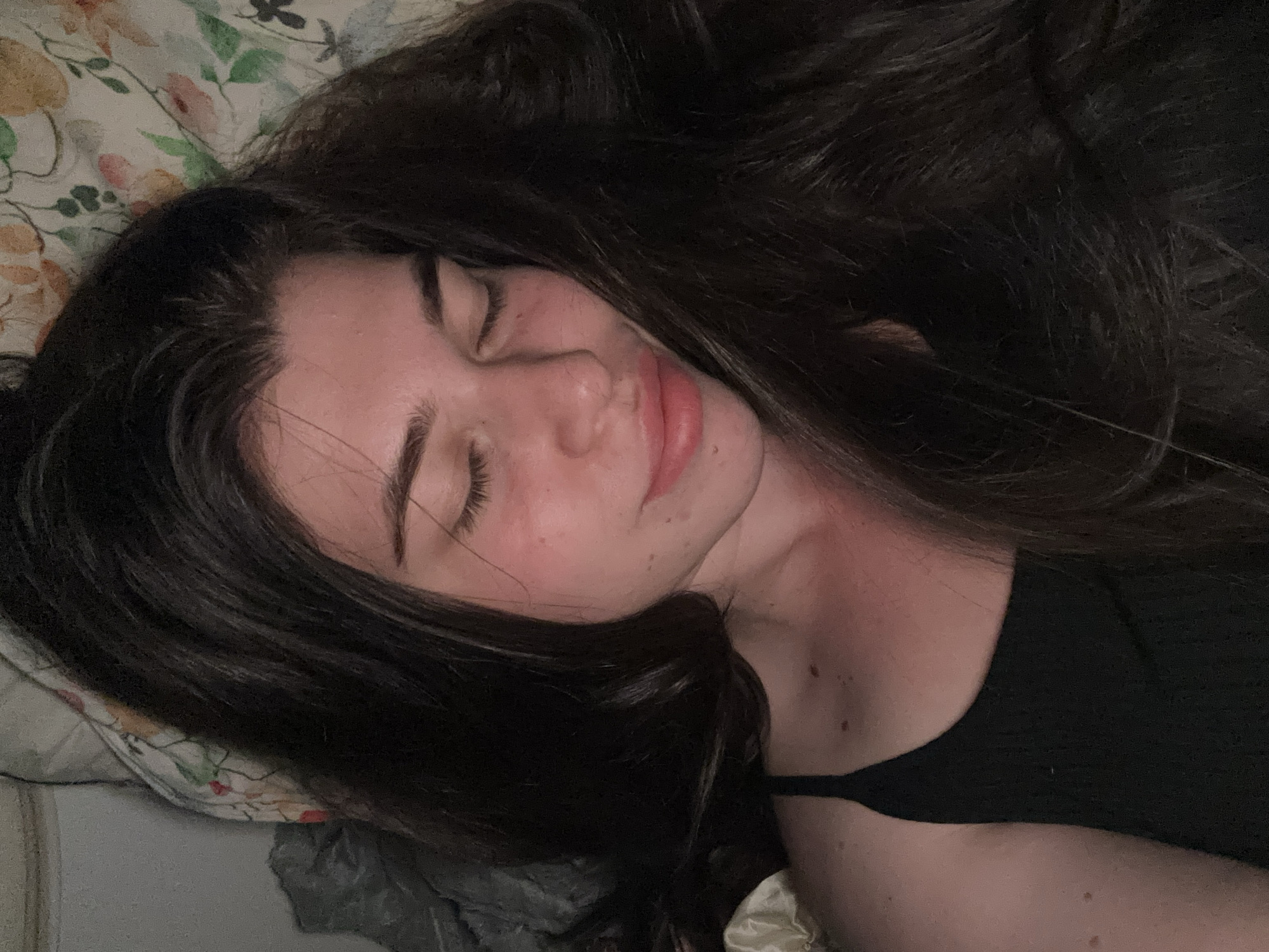A young woman with long, dark hair is lying down on a pillow with a floral pattern. Her eyes are closed. She is wearing a dark-colored top. No elements of the city of Мещёры, Russia are visible in the image.