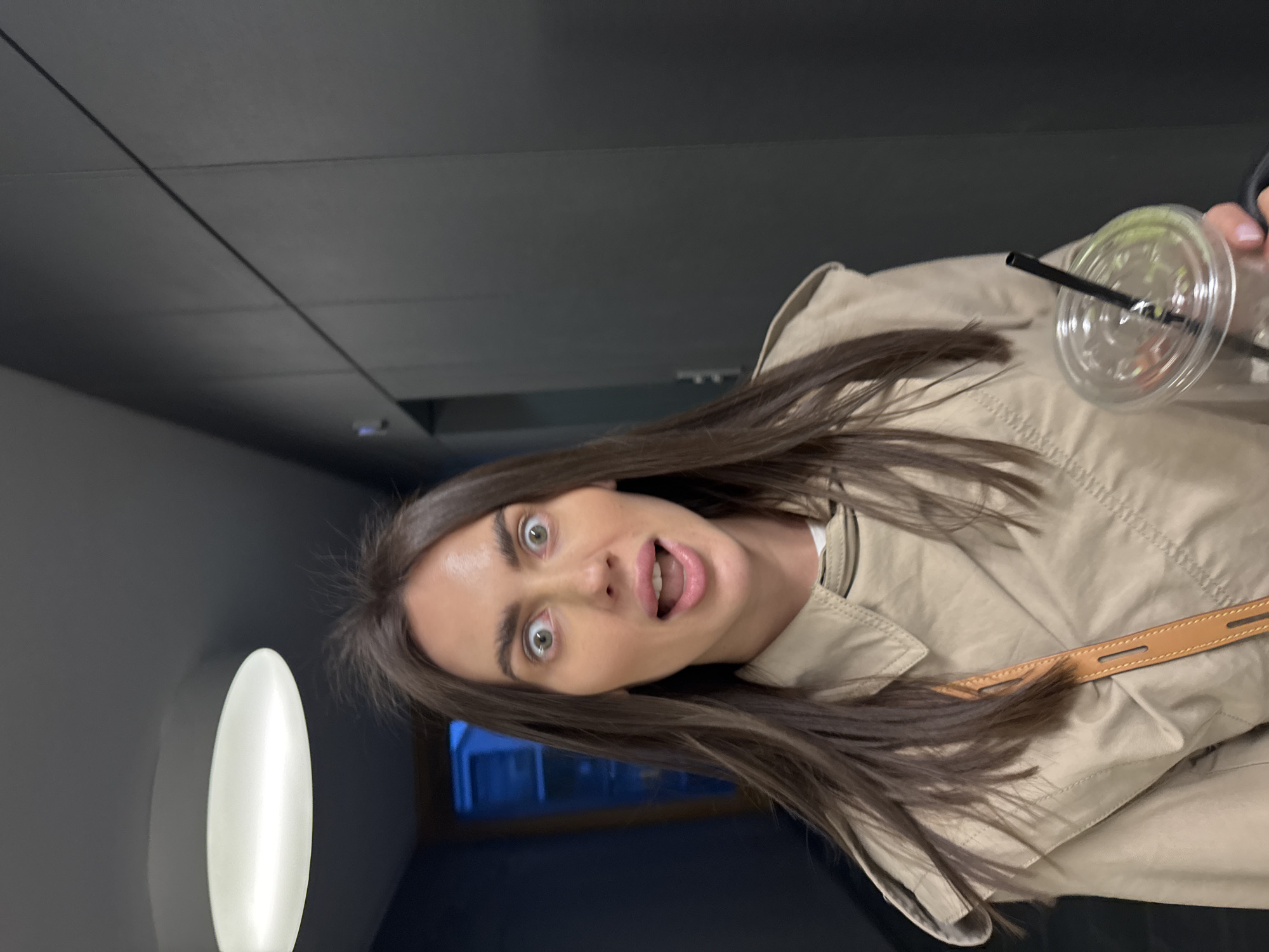 A woman is captured in a selfie-style image, holding a plastic cup with a straw, possibly containing a beverage. She is wearing a light beige trench coat. The background suggests an indoor setting, possibly a hallway or corridor, with a dark gray wall. A distinctive oval-shaped light fixture is visible on the left side of the frame. Through a window, a glimpse of an external environment with blue hues can be seen. The location could potentially be in Warsaw, Poland, as requested.