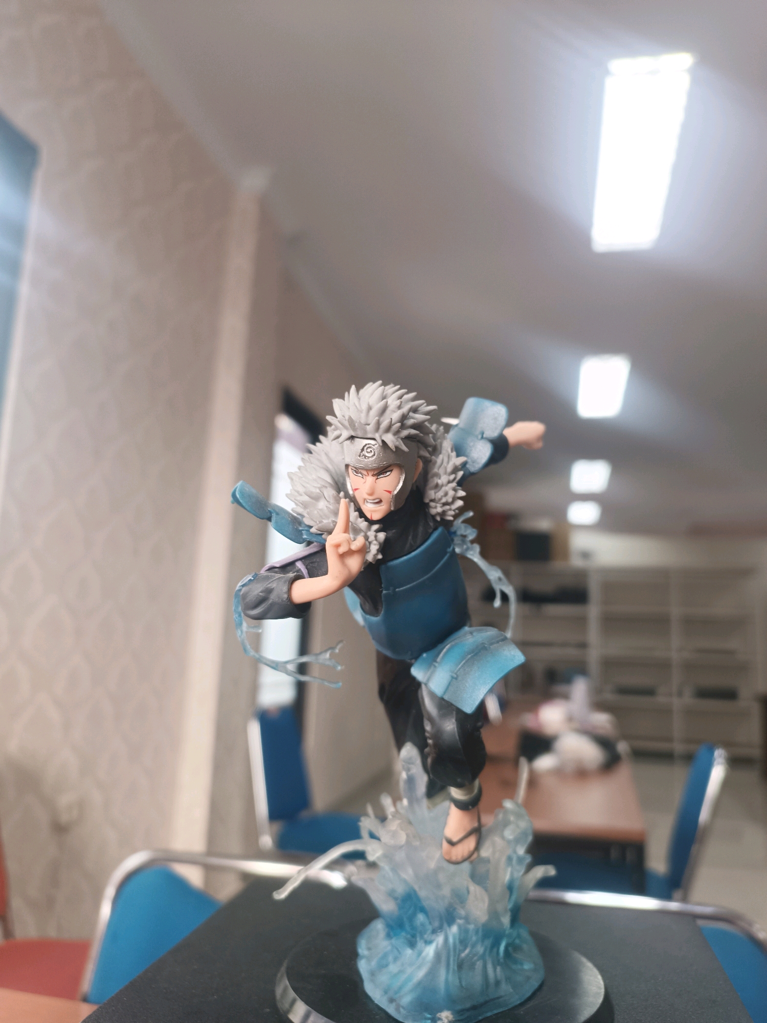 A polyvinyl chloride action figure of Tobirama Senju from the anime series Naruto is displayed on a black surface. The figure is posed in a dynamic stance, with one hand raised and fingers splayed, as if performing a jutsu. It is depicted with its signature white spiky hair, a headband with the Konohagakure symbol, and blue armor over a dark outfit. The figure stands on a translucent blue base designed to resemble water or ice. The background is an indoor environment, possibly an office or a room, with light-colored walls and overhead fluorescent lighting. A blurred table and chairs are visible in the background, along with shelves. The scene is illuminated by artificial light, creating some reflections on the ceiling. The image is taken from a slightly low angle, focusing on the action figure. The location of Tabanan, Indonesia is not discernible from the visual information provided in the image.