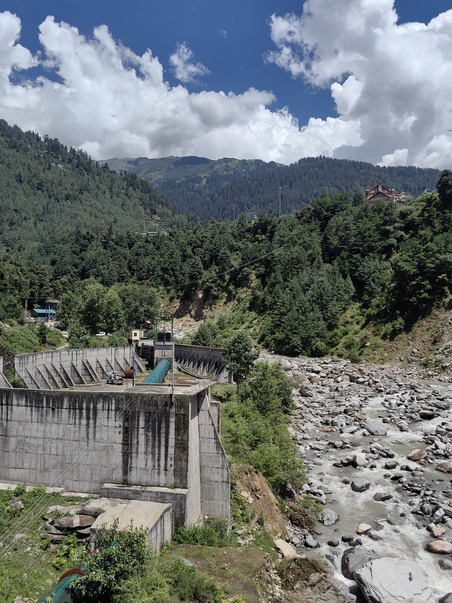 The image captures a scene in Siumsa, India, featuring a concrete dam structure in the foreground. The dam is a substantial, multi-tiered construction with sloped buttresses on its face. Adjacent to the dam, a rocky riverbed is visible with water flowing over the stones. Lush green trees cover the steep hillsides that rise behind the dam and river. The sky is a vibrant blue, dotted with large, fluffy white clouds. In the background, more hills, densely forested, stretch into the distance, with a few buildings visible nestled among the trees on one of the higher slopes. The overall impression is one of a natural landscape complemented by man-made infrastructure.