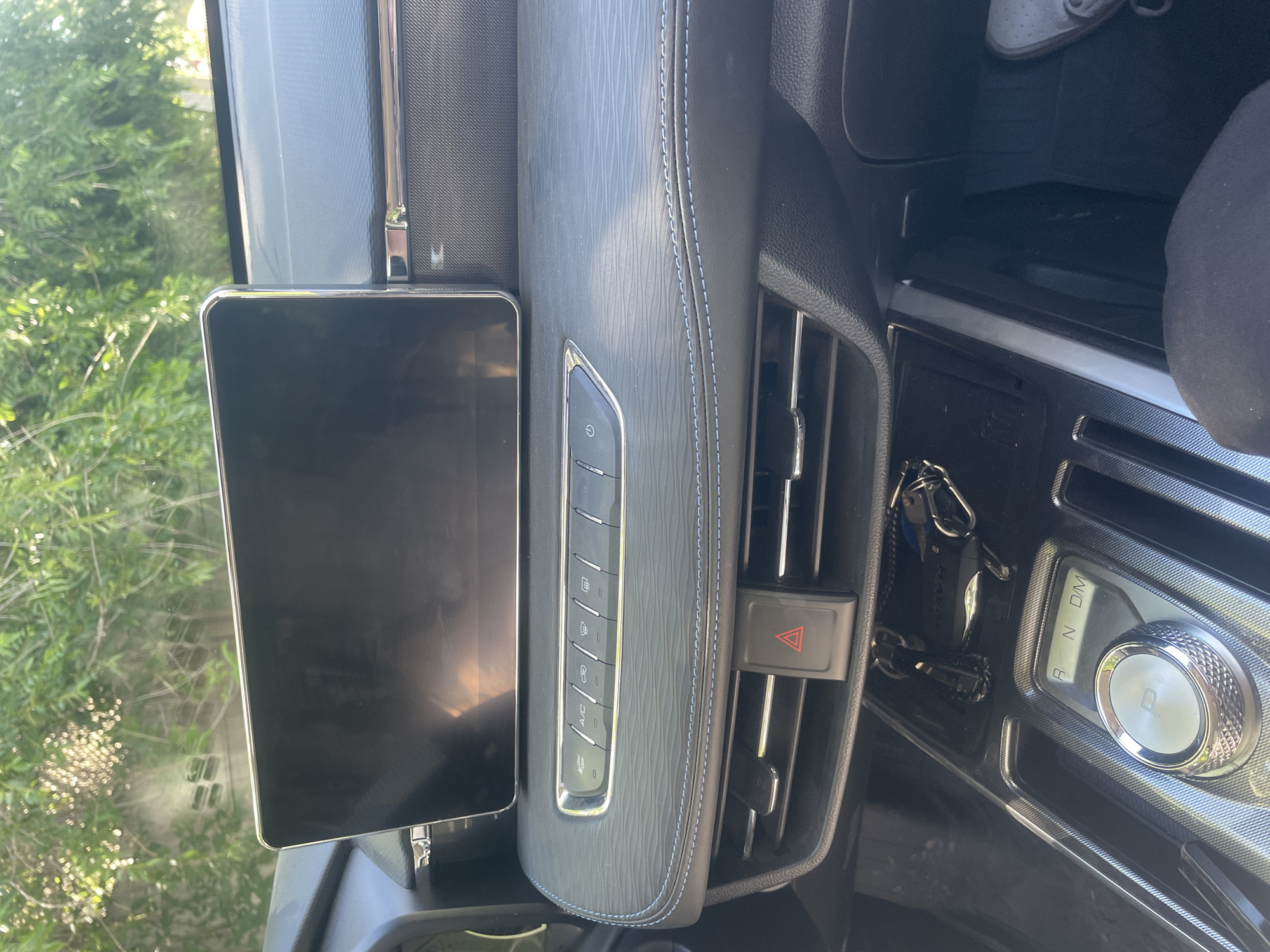 The image shows the interior of a car, likely a modern SUV, with a large central infotainment screen. Below the screen are a series of touch-sensitive buttons for climate control, including icons for A/C, air circulation, defrost, and fan speed. A red hazard light button is prominently placed between two air vents. The gear shifter is a rotary dial with illuminated indicators for Park (P), Reverse (R), Neutral (N), and Drive (D). A set of car keys are hanging from a hook near the shifter. The dashboard is textured with a patterned material and features blue stitching along a seam. The exterior environment, visible through the windshield, consists of green foliage and dappled sunlight, suggesting an outdoor, possibly daytime setting. The image was taken from the driver's perspective.