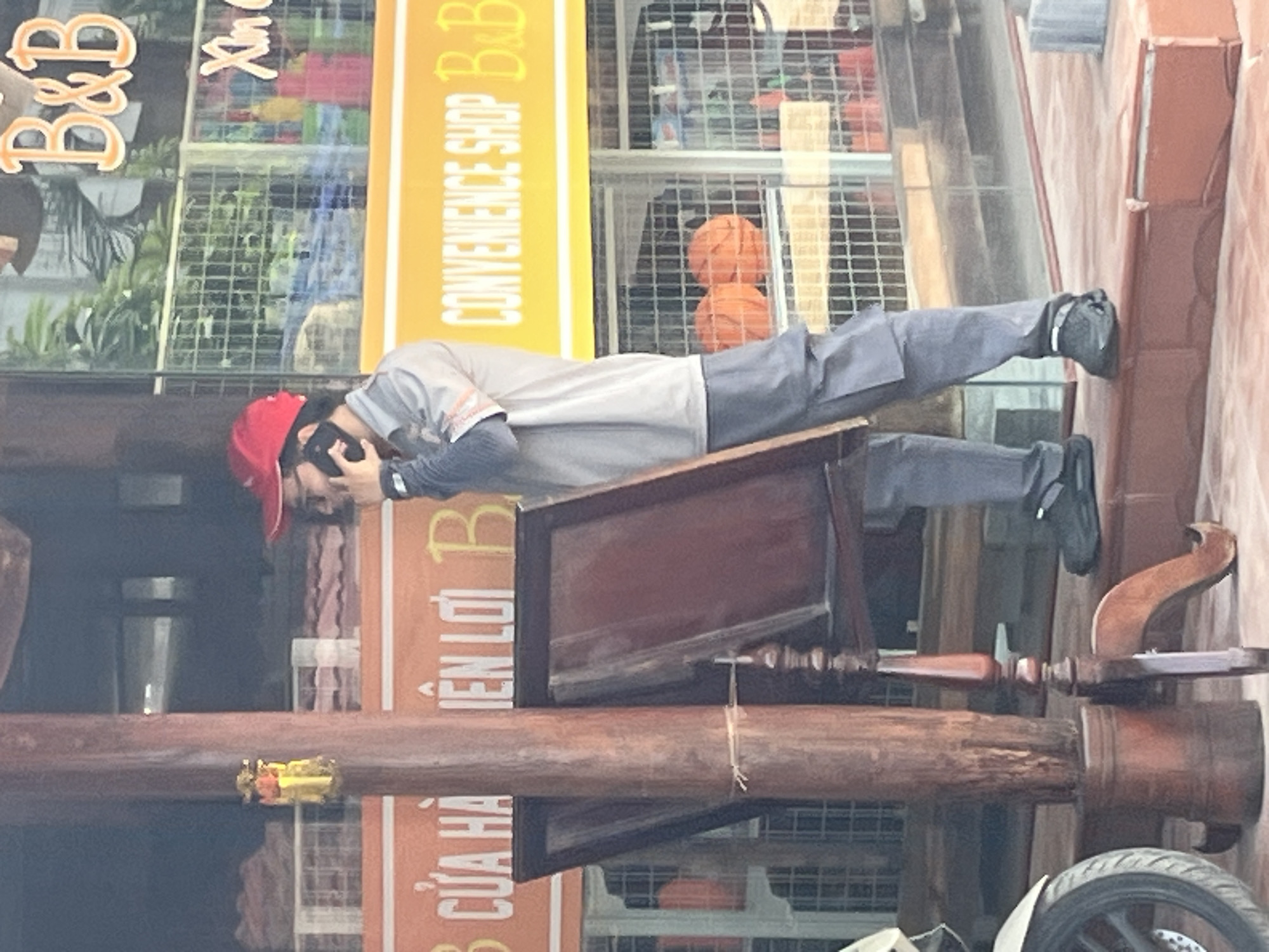 A person wearing a red cap and a gray uniform stands near a wooden pillar and a display stand, holding a black cell phone to their ear. The background features a store with a "B&B" logo and "CONVENIENCE SHOP" text, visible through glass windows. Two orange basketballs are visible inside the store, behind a metal grate. The scene appears to be outdoors in a public space, possibly a street or storefront in Mũi Né, Vietnam, given the store signage.