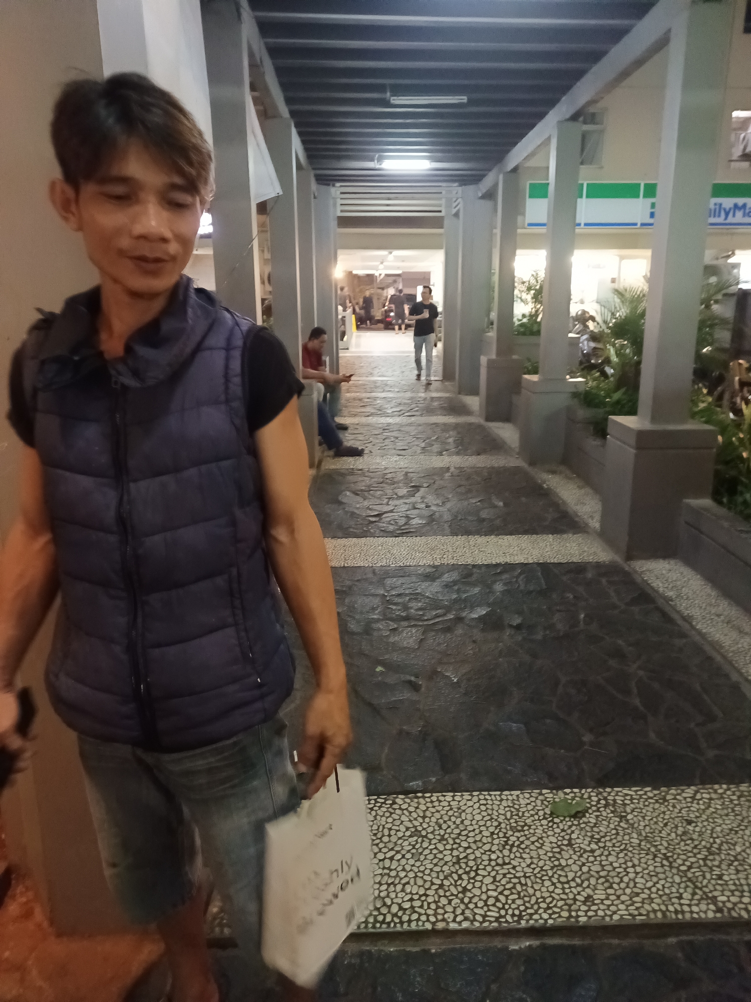 A man wearing a dark blue puffer vest over a black t-shirt and denim shorts stands in a covered walkway, holding a white paper bag in his right hand. The walkway has a patterned floor of dark and light grey stone with a band of small white pebbles. In the background, other people are visible walking along the walkway. To the right, a store with a green and white sign reading "FamilyMart" is visible. The overall scene suggests an outdoor public space, potentially a commercial area, in South Jakarta, Indonesia.