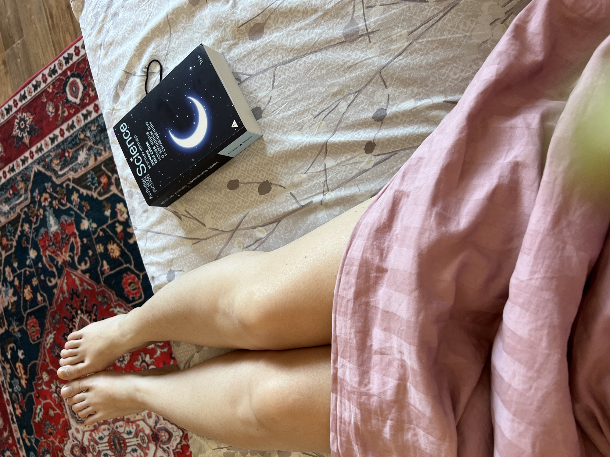 The image shows a close-up, overhead view of a person's legs and feet lying on a bed. The person's feet are bare and resting on a red and black patterned rug. The legs are covered from the knees down with a pink, striped blanket. To the right of the legs, a black book titled "Science" with a crescent moon on the cover is placed on a white and grey patterned sheet. The book is marked with "18+". The overall scene suggests a relaxed, indoor setting. The specific location in the city of Novosibirsk, Russia cannot be determined from the image.