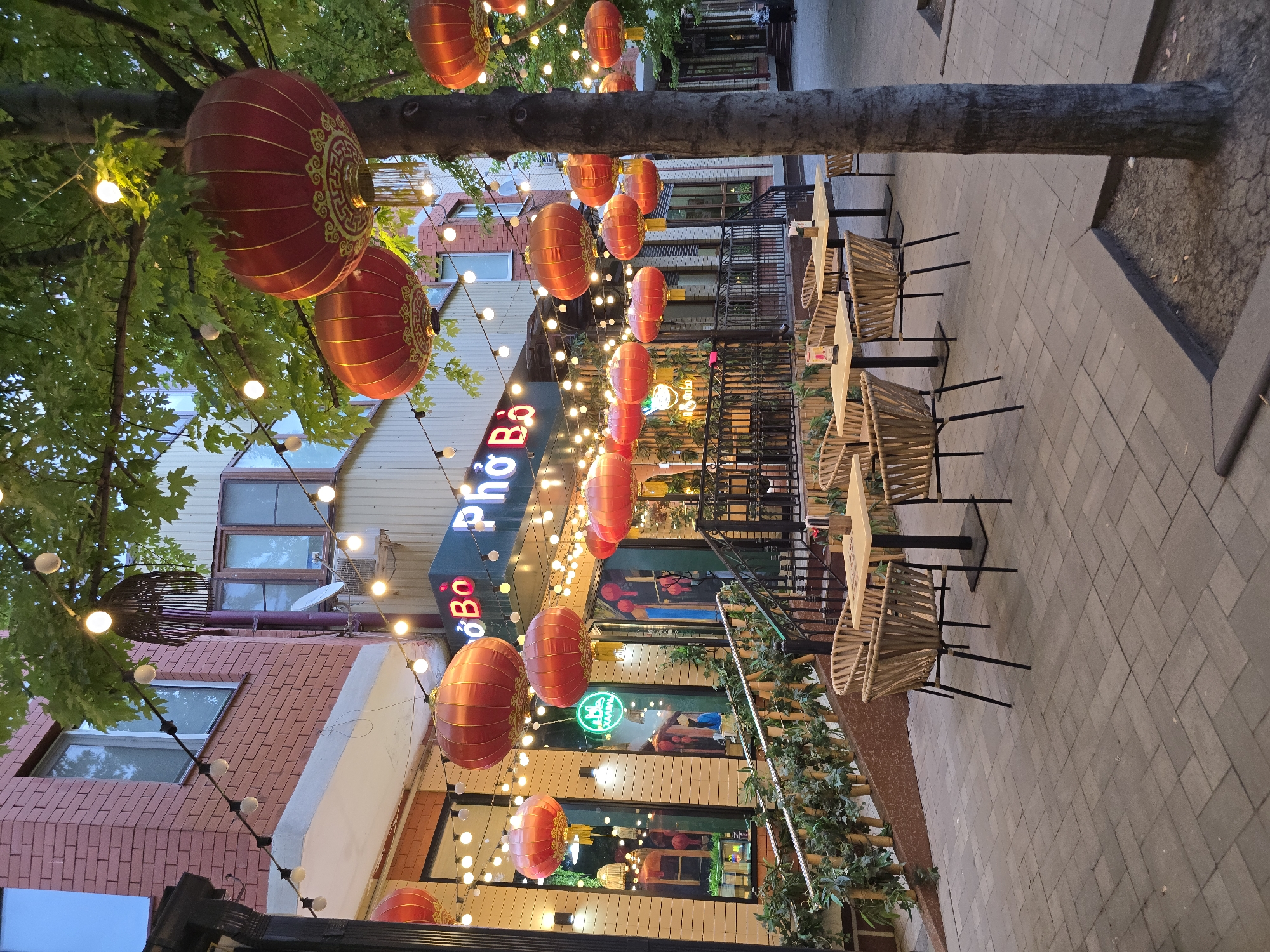 The scene depicts an outdoor dining area of a restaurant, likely in an urban setting. The restaurant, named "Pho Bo," is adorned with red lanterns and string lights, creating a warm and inviting ambiance. Several tables and chairs are set up outside, suggesting it is open for service. The surrounding environment includes brick buildings and trees, adding to the urban landscape. The time of day appears to be evening or dusk, given the presence of artificial lighting.