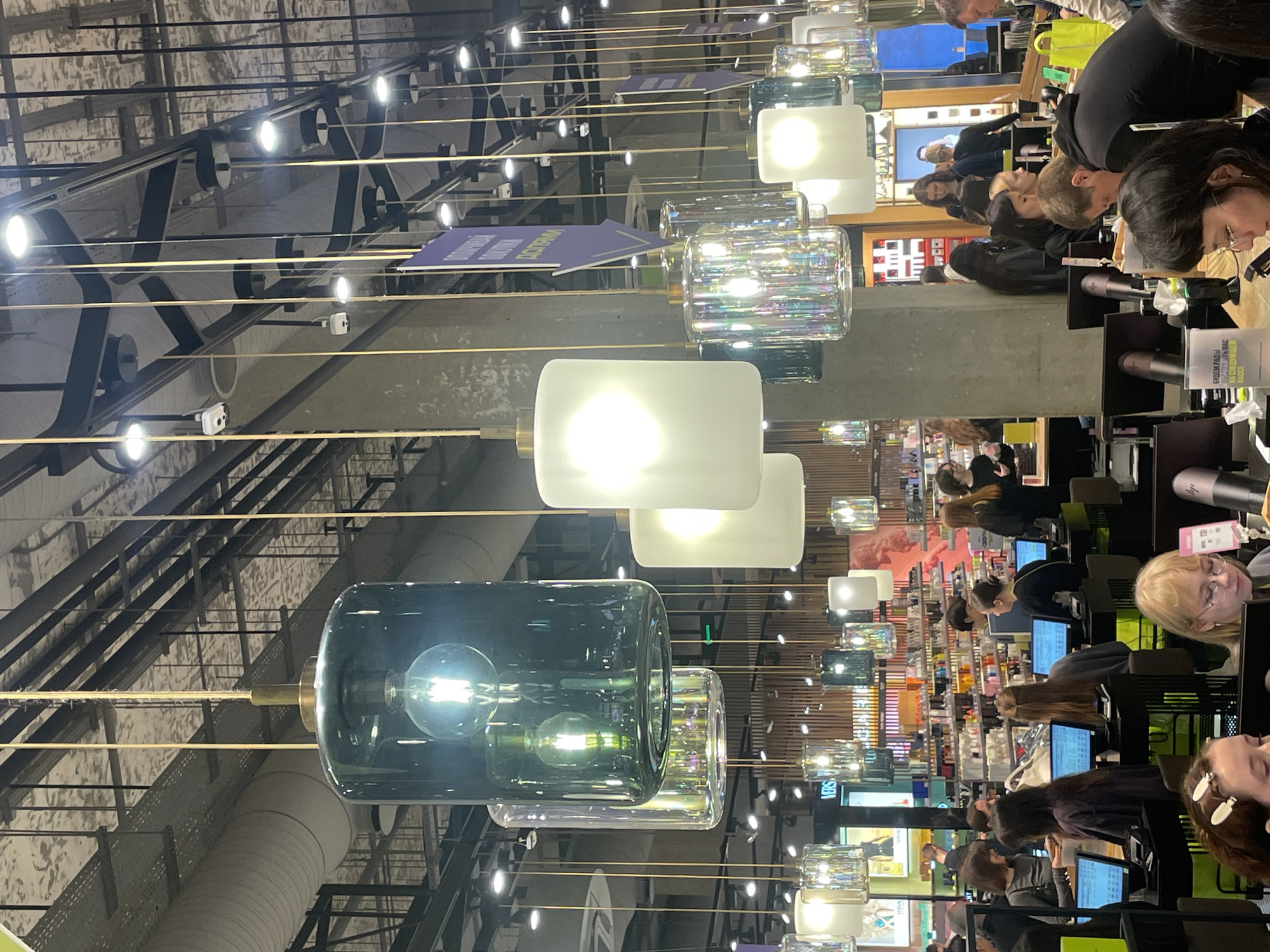 The image shows a brightly lit commercial space, possibly a cosmetics or perfume department within a larger store. A row of people are seated at what appear to be testing stations or makeup counters, each with a small monitor. The people are primarily female and seem to be engaged in trying out products or receiving consultations.
Above, a complex network of industrial-style metal beams and pipes forms the ceiling structure. Suspended from this structure are a series of decorative pendant lights with cylindrical or rectangular shades in various materials, including colored glass.
Based on the Cyrillic text visible on signage, specifically the phrase "БУДЕМ РАДЫ ОБСЛУЖИТЬ ВАС НА СЛЕДУЮЩЕМ КАССЕ", it is likely that this photo was taken in a store in Russia. Given the architectural style and the merchandise, it could be located in a modern shopping mall or department store in a major Russian city, such as Saint Petersburg.