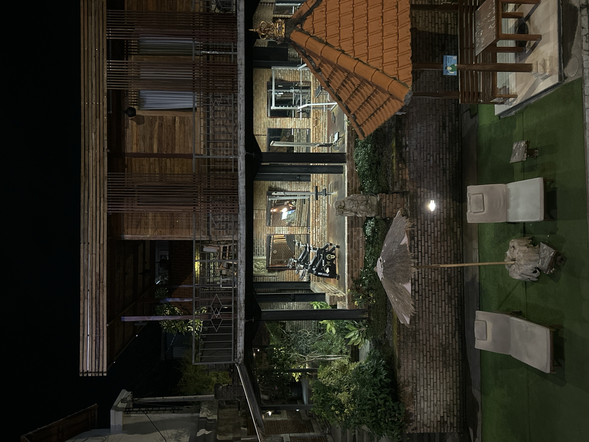 The image shows an outdoor scene at night. In the foreground, there are two light-colored lounge chairs on a green surface, possibly artificial turf. To the right of the lounge chairs is a small, rustic thatched umbrella and a stone base. Further to the right, there is a wooden gazebo with a terracotta tiled roof. Inside the gazebo, there is a wooden bench and a wooden table.
In the mid-ground, there is a brick wall that extends across the width of the image. Above the brick wall, there is a building with multiple levels constructed primarily of wood. The first floor of the wooden building has a covered walkway or patio area with several exercise machines, including spinning bikes and weight-lifting equipment, visible through open walls. There are also potted plants and trees in this area. The second floor of the wooden building features balconies with railings and wooden slatted walls.
In the background, there is a glimpse of another building with a white facade. The overall environment appears to be a resort or hotel complex, likely in a tropical or sub-tropical location given the style of architecture and vegetation. The lighting suggests it is nighttime, with artificial lights illuminating the area. The location is identified as North Kuta, Indonesia.