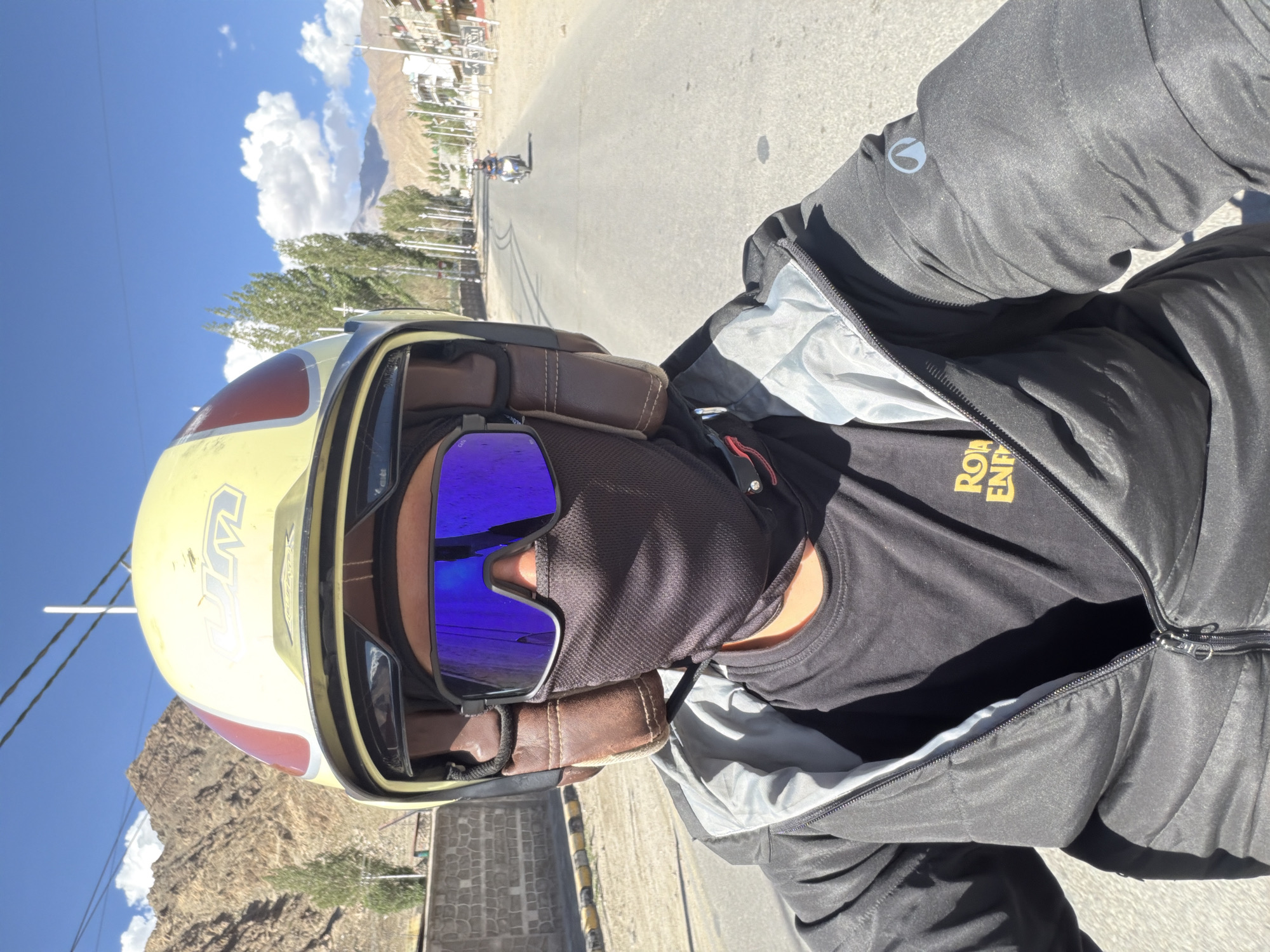 A close-up selfie shows a person riding a motorcycle on a road in Leh, India. The rider wears a yellow and red helmet, blue mirrored goggles, and a black face mask. They are dressed in a black t-shirt with a yellow logo and a black puffer jacket. The road stretches into the distance, with buildings and trees lining the sides. Mountains are visible in the background under a blue sky with scattered clouds. Another motorcycle is visible further down the road.