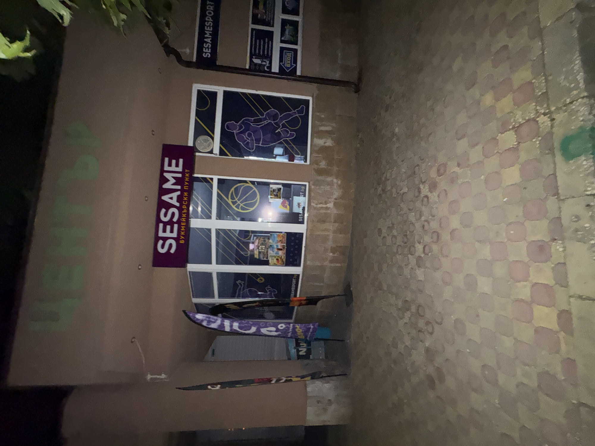 The image shows a storefront at night in Stamboliyski, Bulgaria. The storefront has large windows displaying graphics of basketball players and a basketball. Above the windows, a sign reads "SESAME" in large letters, and below it, smaller text in Cyrillic reads "БУКМЕЙКЪРСКИ ПУНКТ" which translates to "BOOKMAKING POINT". To the right of the main storefront, another sign reads "SESAMESPORT.BG". There are also several flags displayed in front of the storefront. The ground in front of the building is paved with interlocking stones, some of which are illuminated by a faint green light. The overall lighting suggests it is nighttime, and the scene is relatively quiet. The upper part of the building shows some graffiti in green letters that appears to spell "ЦЕНТЪР", meaning "CENTER".