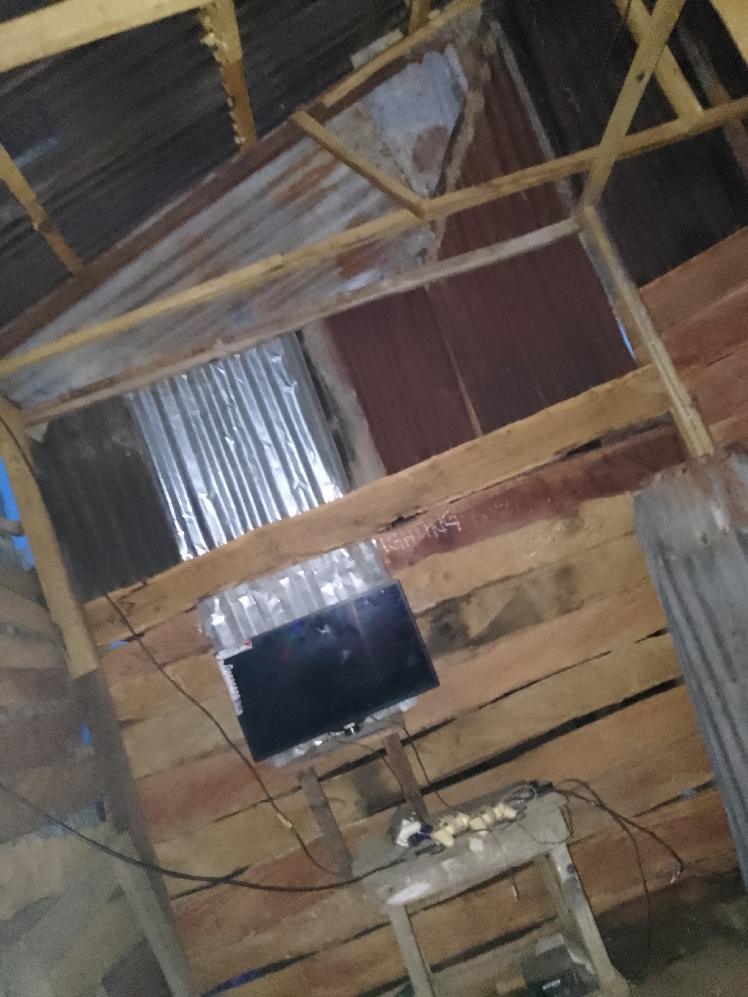 The image depicts the interior of a rudimentary structure, likely a dwelling, constructed with unpainted wooden planks for walls and a corrugated metal roof. The overall impression is one of simple, possibly temporary housing. There is a black television set perched on a makeshift stand constructed from wood and possibly concrete blocks. Various cables are visible, connecting to the television and other unseen electronics. The walls are rough-hewn, showing visible grain and knots in the wood, with some signs of age and wear. The corrugated metal roofing is exposed, with some sections appearing rusted or stained. In the background, more corrugated metal sheeting is visible, possibly used as a partition or as part of the external structure. A partial inscription, "NGIDING INS" is faintly visible in white lettering on one of the wooden planks. There is no visible evidence of people in the image. The lighting appears dim, suggesting it may be late evening or the interior is not well-lit. The scene is consistent with informal settlements or rural housing found in Serti, Nigeria, characterized by the use of locally sourced and readily available materials for construction.