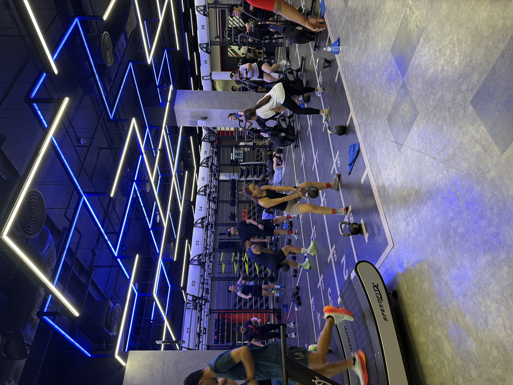 The image depicts the interior of a modern gym, likely located in Nong Pa Khrang, Thailand, characterized by its industrial aesthetic with exposed ceilings and a dynamic lighting system featuring blue and yellow LED strips. A group of individuals are engaged in various fitness activities. In the foreground, a person is running on a treadmill. To the right, several people are performing exercises with kettlebells. In the mid-ground, individuals are using stationary bikes, and others are observing or resting. Equipment such as weight racks, mats, and exercise machines are visible throughout the space, all contributing to a functional and active gym environment.