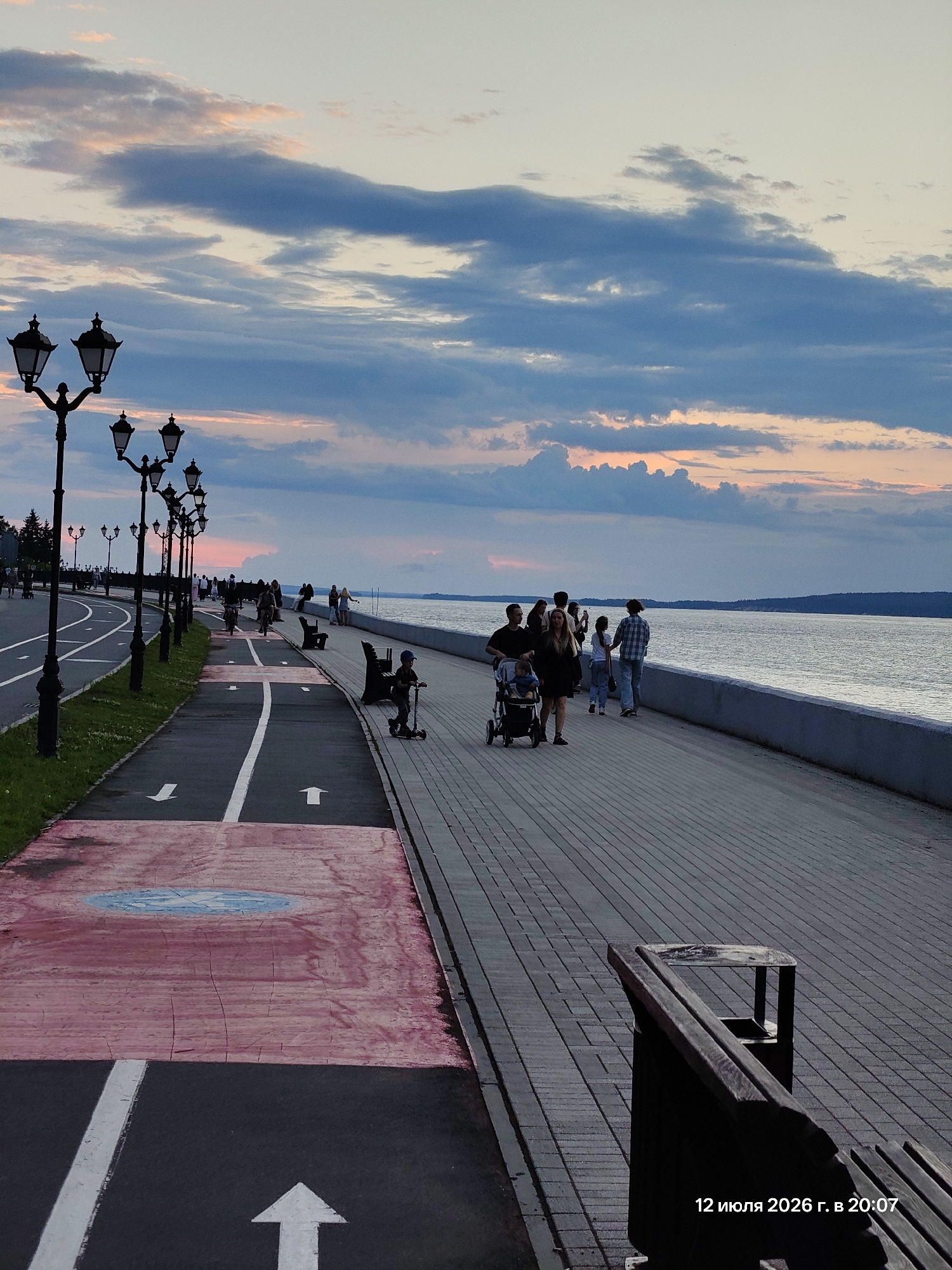 The image depicts a waterfront promenade at dusk. A paved walkway runs parallel to a body of water, with a designated bike lane and a sidewalk for pedestrians. Several lampposts line the promenade, casting a warm glow. A group of people are seen strolling along the walkway, some with a stroller. A child is riding a scooter on the bike lane, and a cyclist is visible in the distance. Benches are placed along the promenade for resting. The sky is filled with colorful clouds, suggesting a sunset or sunrise. In the bottom right corner, a timestamp indicates "12 июля 2026 г. в 20:07", which translates to "July 12, 2026 at 8:07 PM".