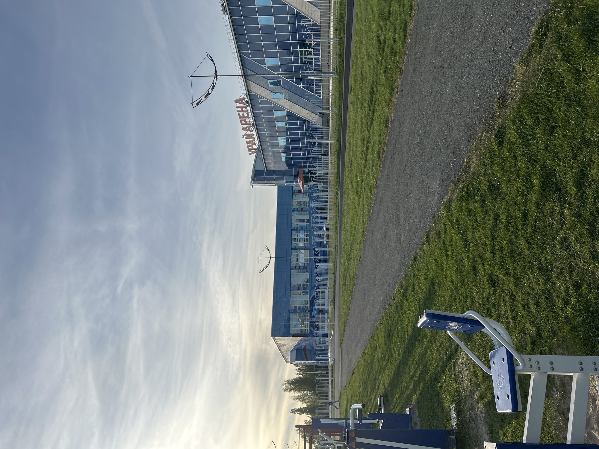 The image shows the Ugra Arena in Uray, Russia. The arena is a large, modern building with blue and silver facades. In front of the arena, there is a paved path and a grassy area with outdoor exercise equipment. The sky is partly cloudy. The sun is low on the horizon.